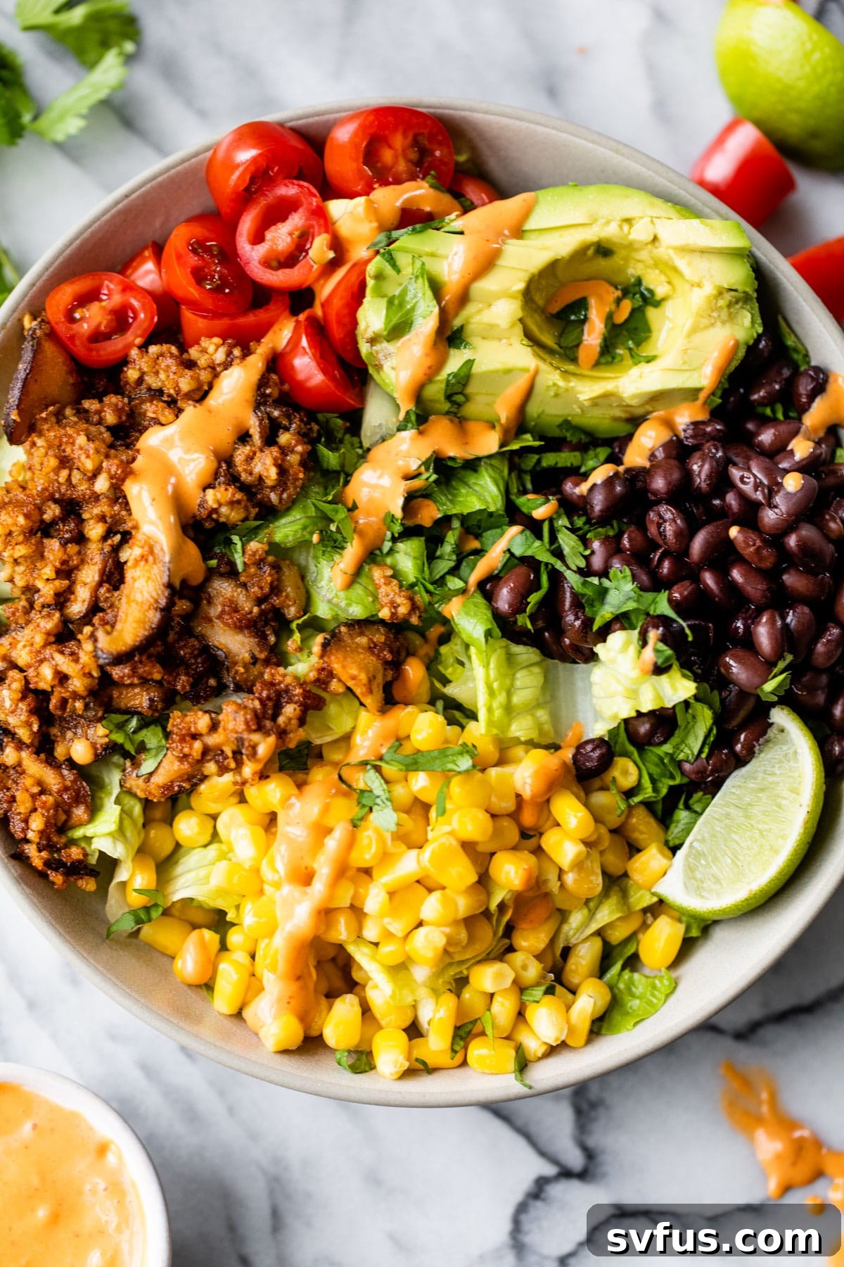 A beautifully presented bowl of Vegan Taco Salad, featuring a generous portion of walnut taco meat nestled on a bed of fresh romaine lettuce, surrounded by vibrant black beans, sweet corn, diced tomatoes, and creamy avocado slices, all drizzled with a zesty chipotle aioli.
