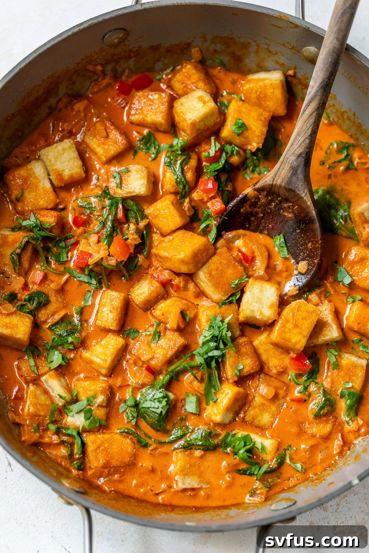 A wooden spoon delicately scooping up a portion of rich tofu curry from a large, inviting grey skillet, highlighting its creamy texture.