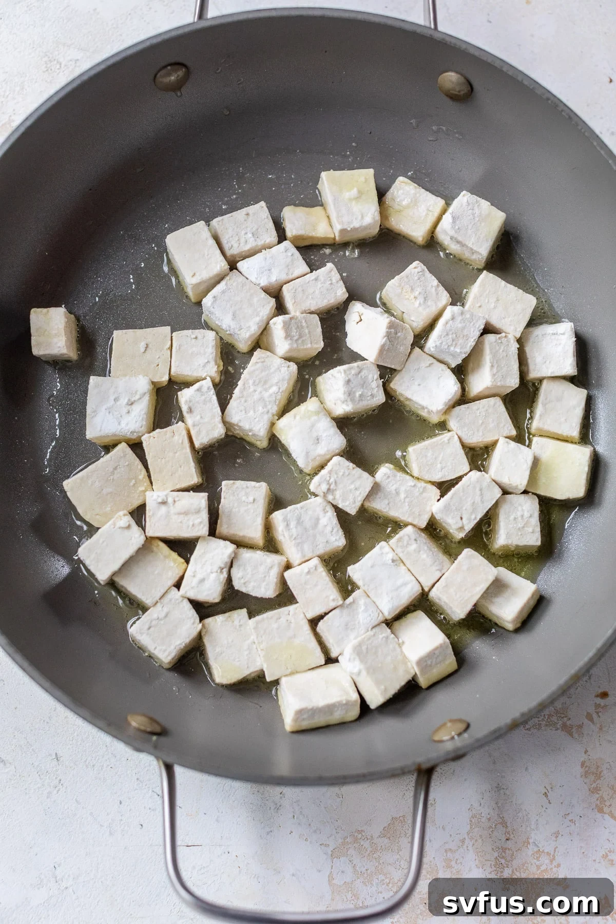 Frying cornstarch-coated tofu cubes in a large grey skillet, showing the initial cooking stage.