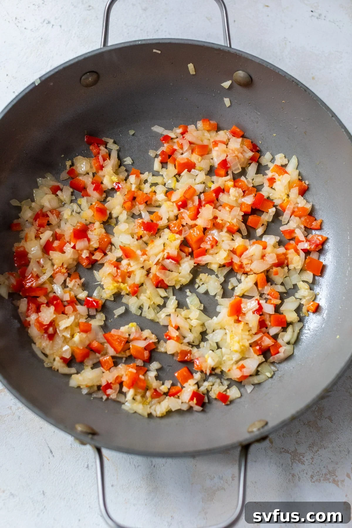 Diced onions, red peppers, and minced garlic being sautéed in a large grey skillet, creating the aromatic base for the curry sauce.