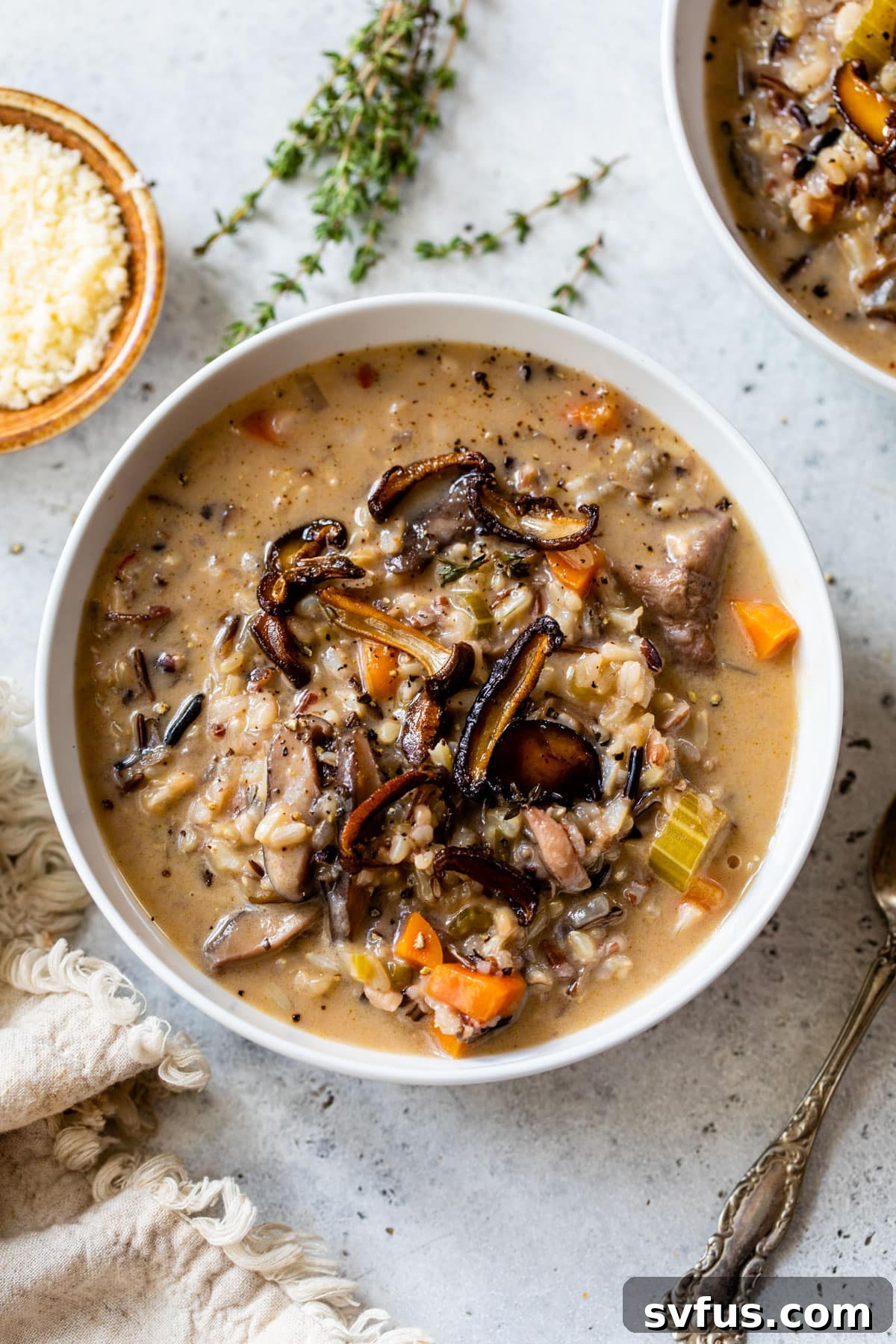 A close-up of a bowl of creamy wild rice and mushroom soup, garnished with fresh thyme, next to a small bowl of grated Parmesan.