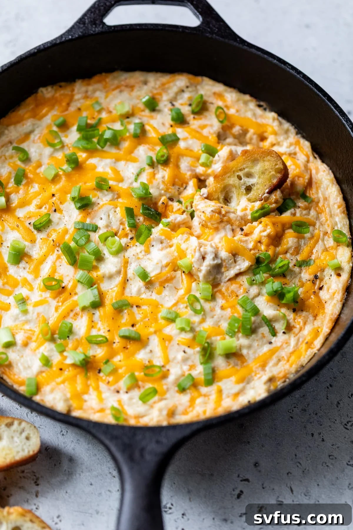 A baguette slice being dipped into a bubbly, golden hot crab dip served in a large black cast iron skillet.