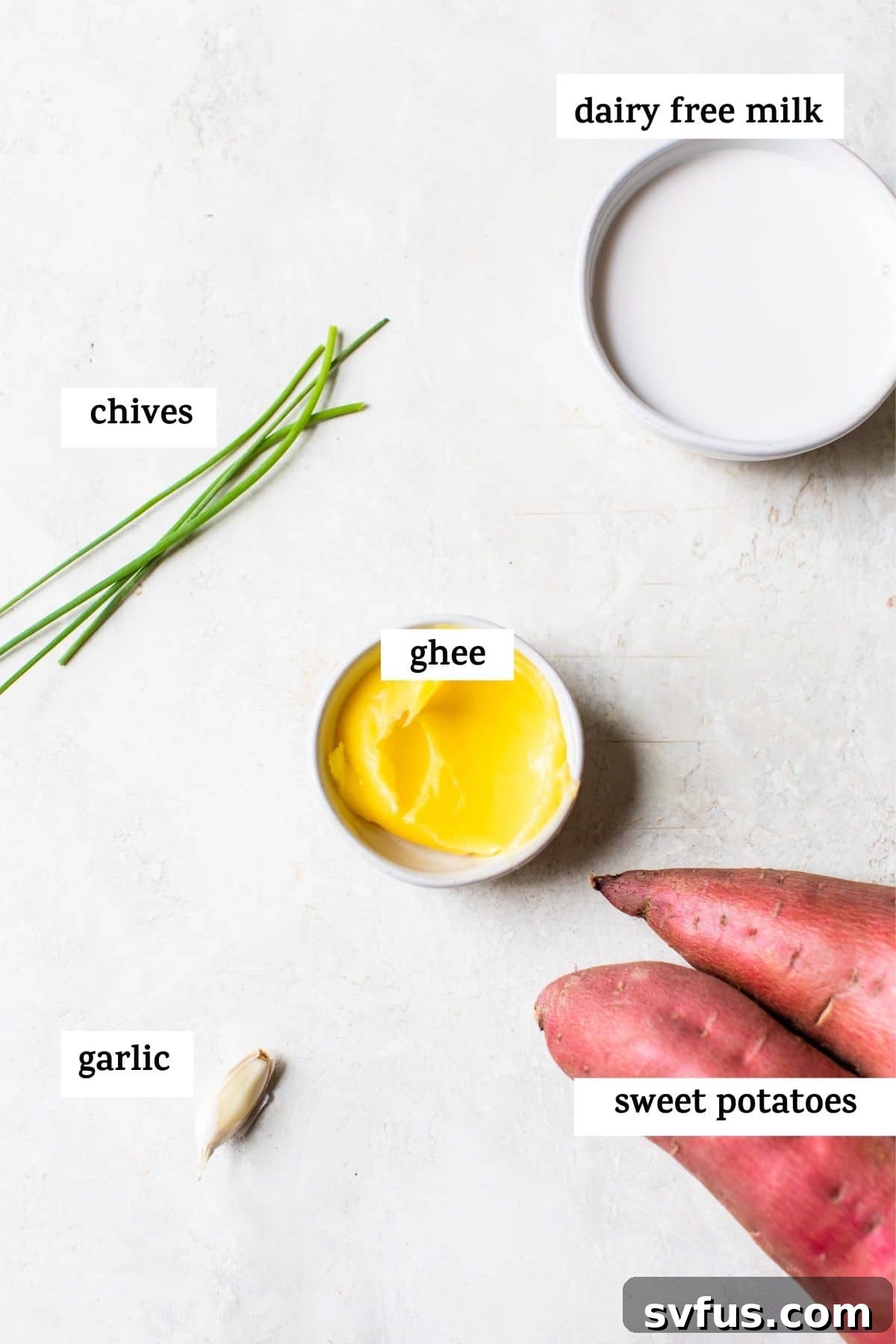 Four essential ingredients for savory mashed sweet potatoes laid out on a rustic wooden table, including whole sweet potatoes, a jar of ghee, fresh garlic cloves, and a carton of almond milk.
