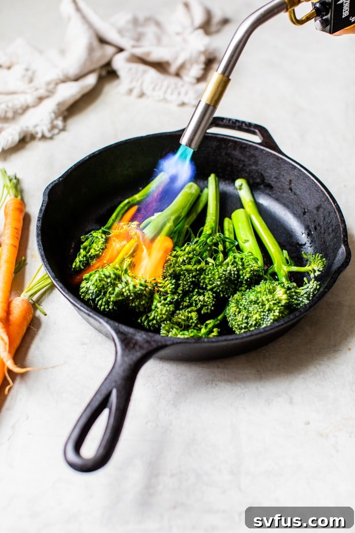 Broccolini being skillfully charred in a cast iron skillet using a kitchen torch, showing the process of achieving perfect smoky edges.