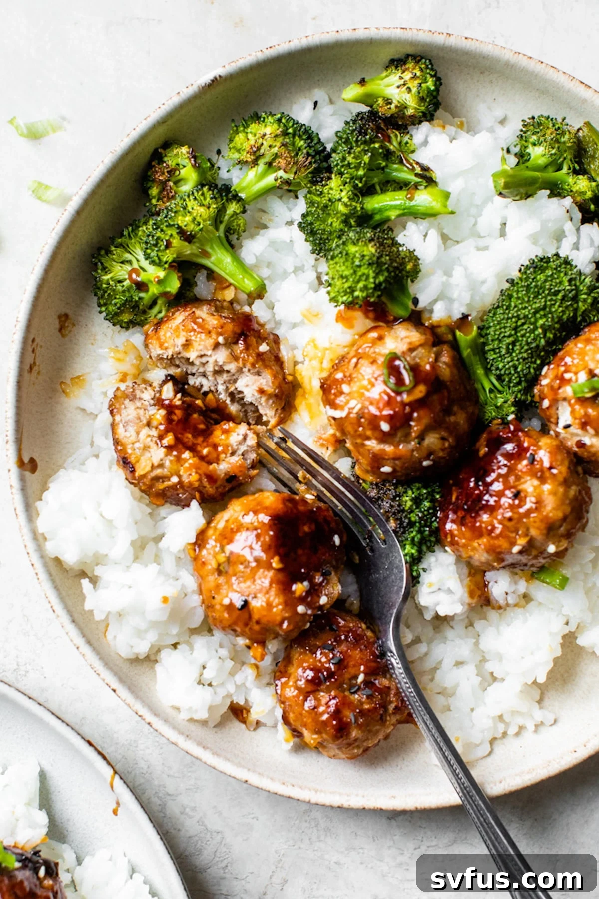 A vibrant bowl of white rice topped with green broccoli florets and saucy teriyaki pork meatballs.