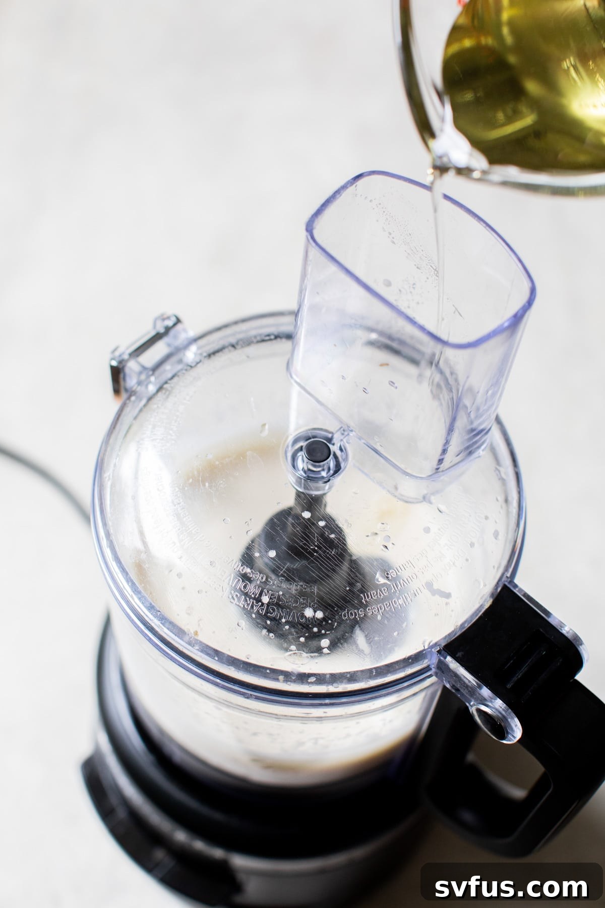 Avocado oil being poured slowly into the top feed tube of a running food processor, emulsifying with other ingredients.