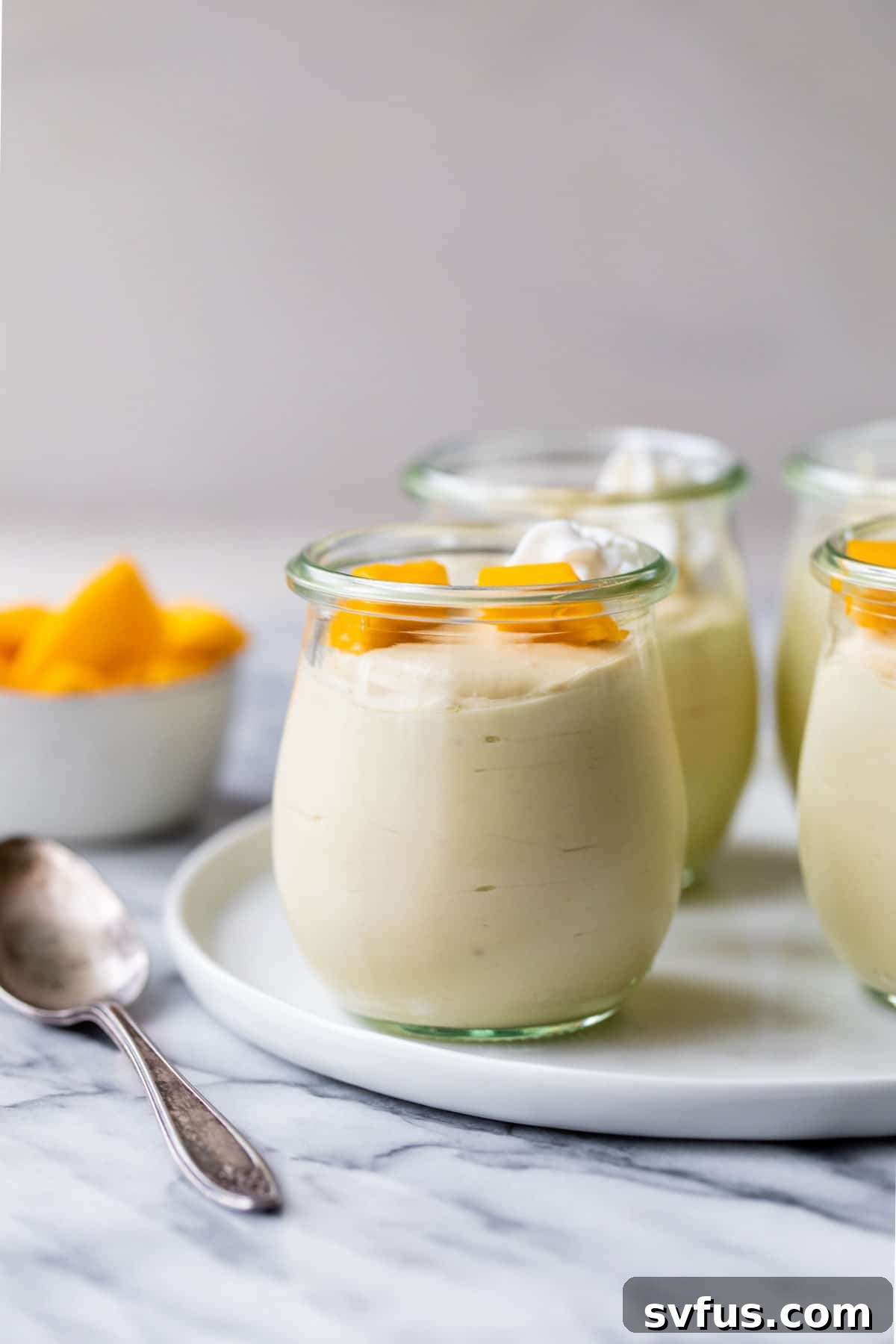 Four elegant jars of vegan mango mousse sit beside a small bowl overflowing with fresh, vibrant mango chunks, ready to be devoured as a refreshing summer treat.