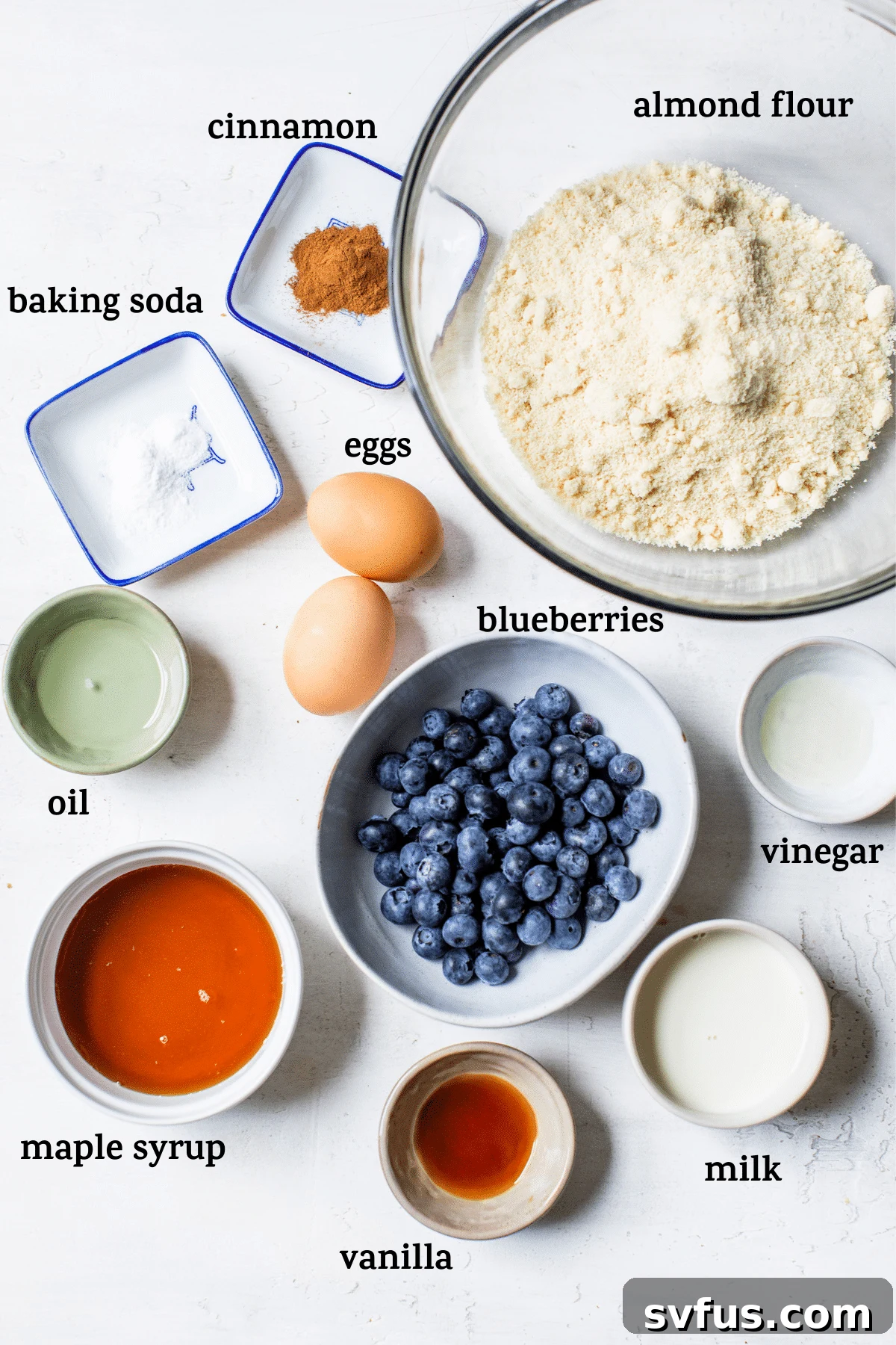A collection of fresh ingredients laid out on a kitchen counter for making blueberry muffins, including almond flour, eggs, maple syrup, blueberries, milk, and spices