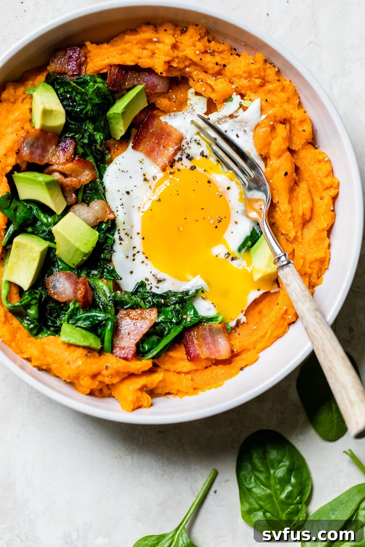 A close-up of the fully assembled sweet potato breakfast bowl, featuring mashed sweet potatoes, crispy bacon, wilted spinach, and a perfectly fried egg with a runny yolk.