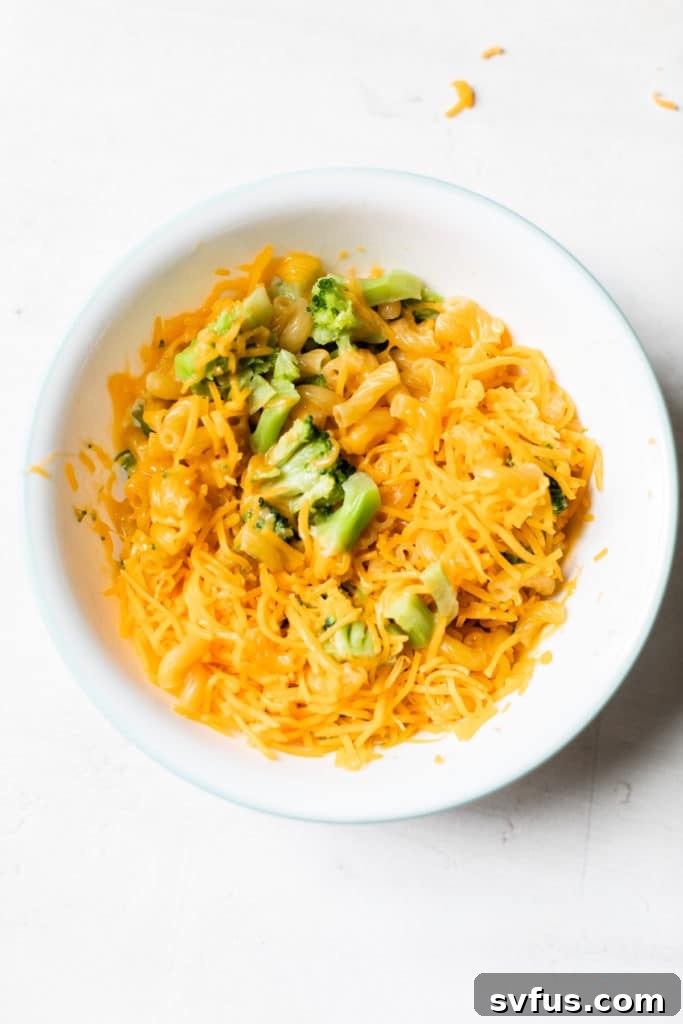A bowl of partially cooked pasta and broccoli, now with shredded cheddar cheese added, ready for the final microwave heating.