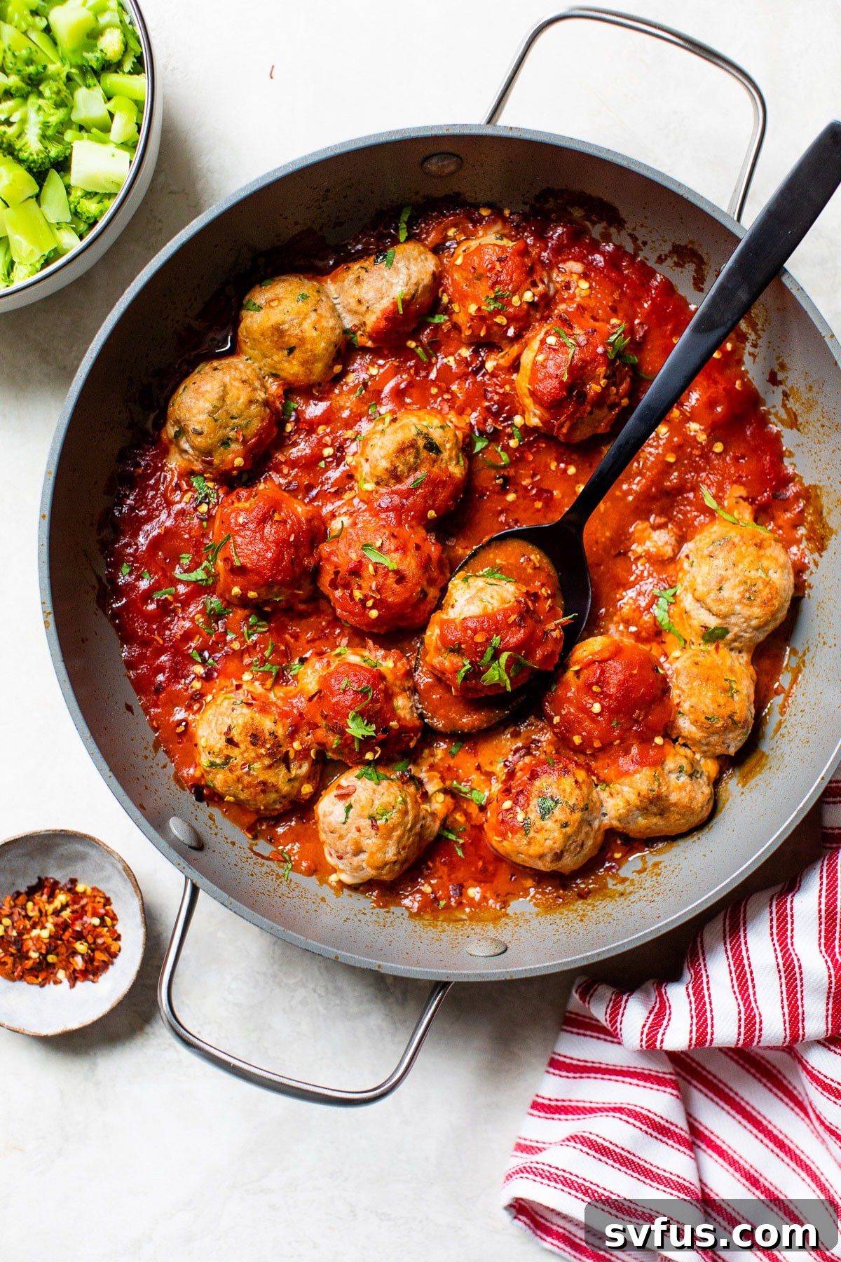 A cast-iron skillet filled with tender paleo turkey meatballs in marinara sauce, with a side of steamed broccoli florets.