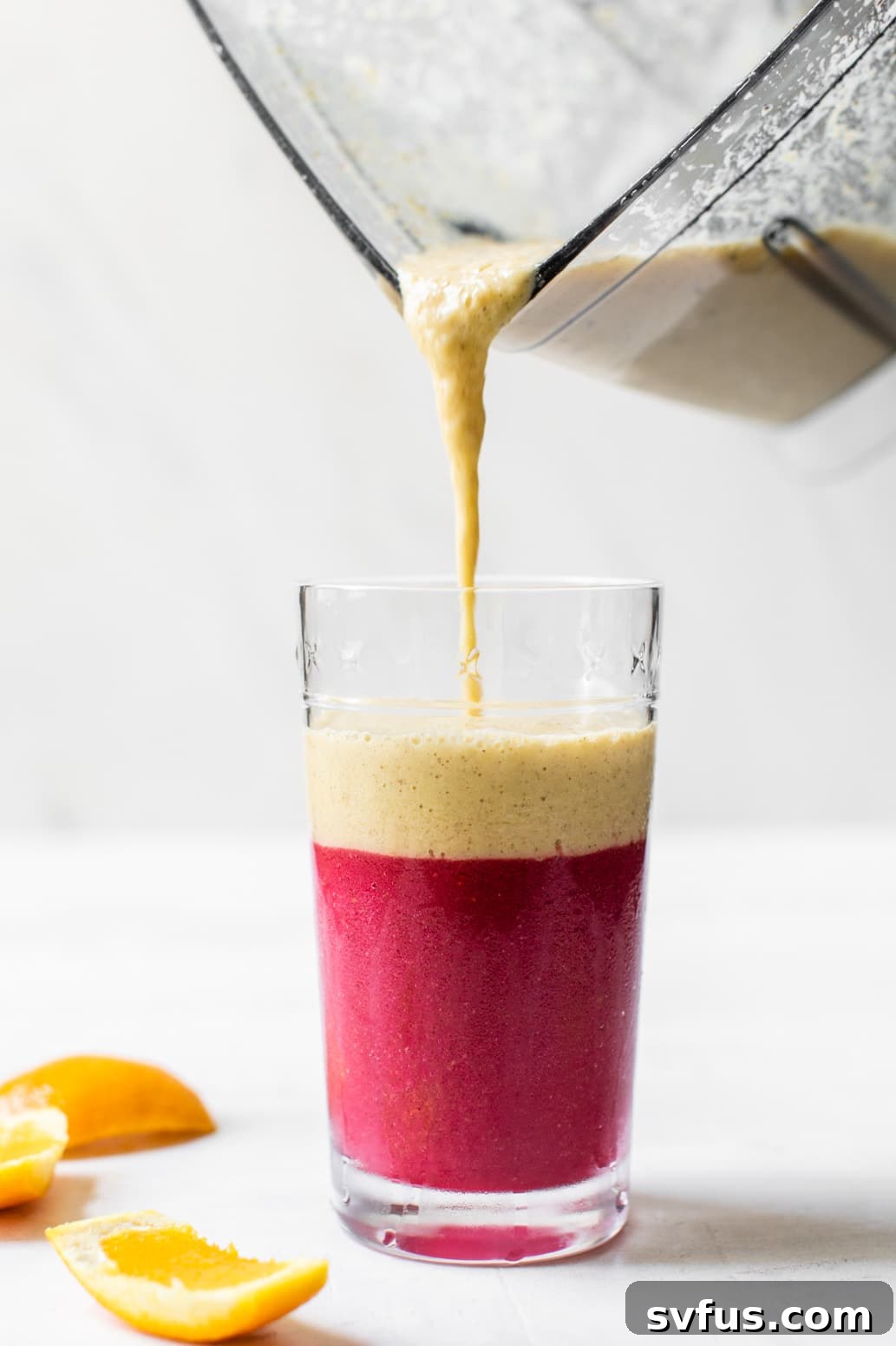 pouring a vibrant orange smoothie from a blender into a glass over a pink raspberry layer