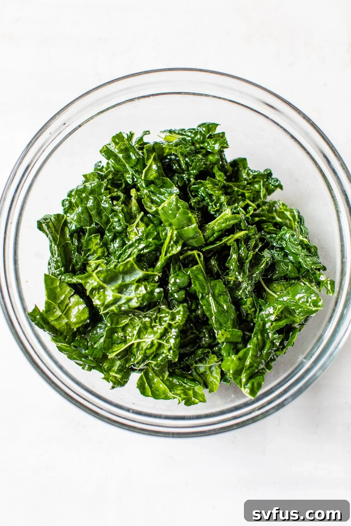 Freshly massaged kale glistening with olive oil in a clear glass bowl, ready for other ingredients.