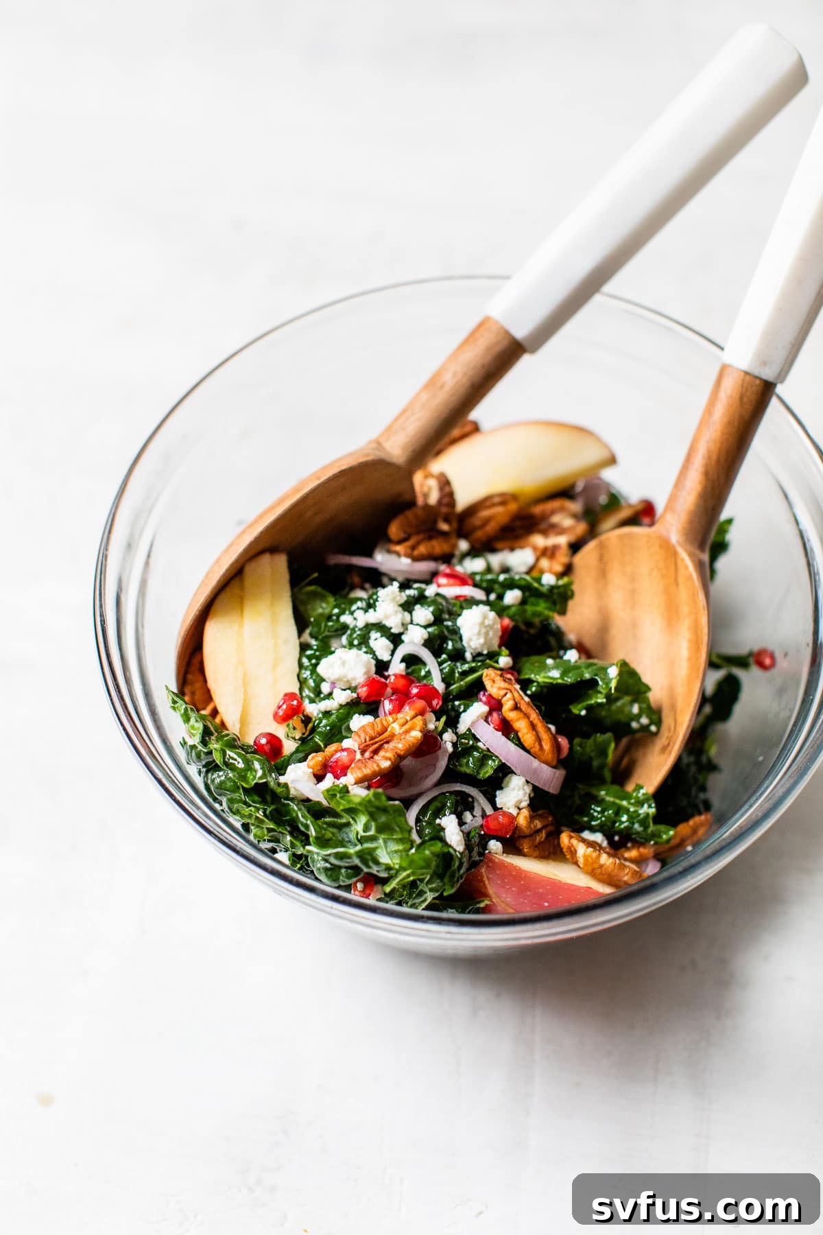 A large bowl of winter kale salad being tossed with kitchen tongs, showing the mix of kale, apple, pecans, and pomegranate.