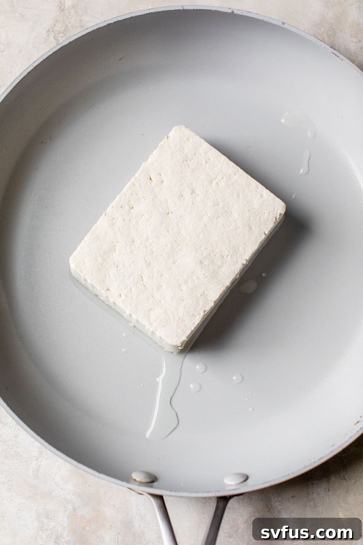 A firm block of tofu resting in a hot skillet, ready to be broken down and transformed into a savory scramble.