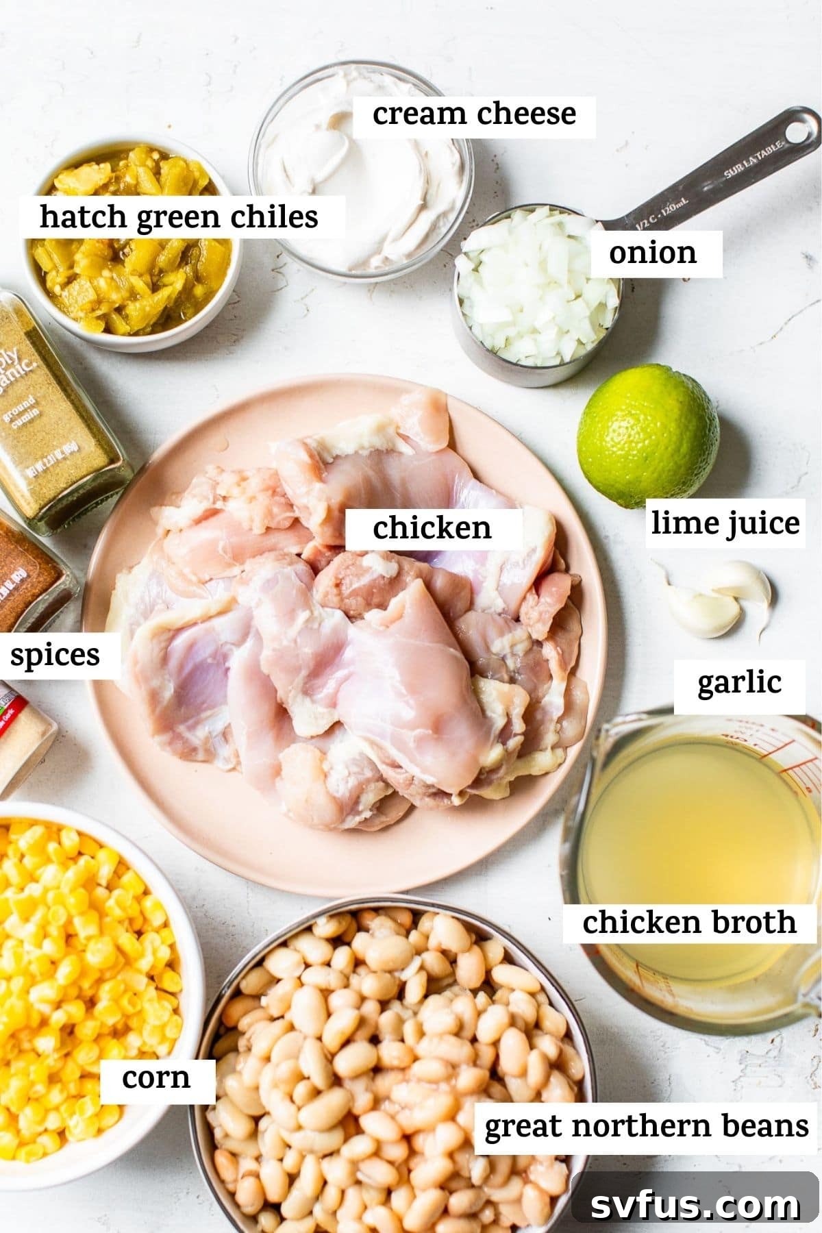 A collection of fresh ingredients for white chicken chili, including canned beans, frozen corn, diced onions, garlic, green chiles, spices, chicken, and cream cheese, arranged in bowls on a wooden table.