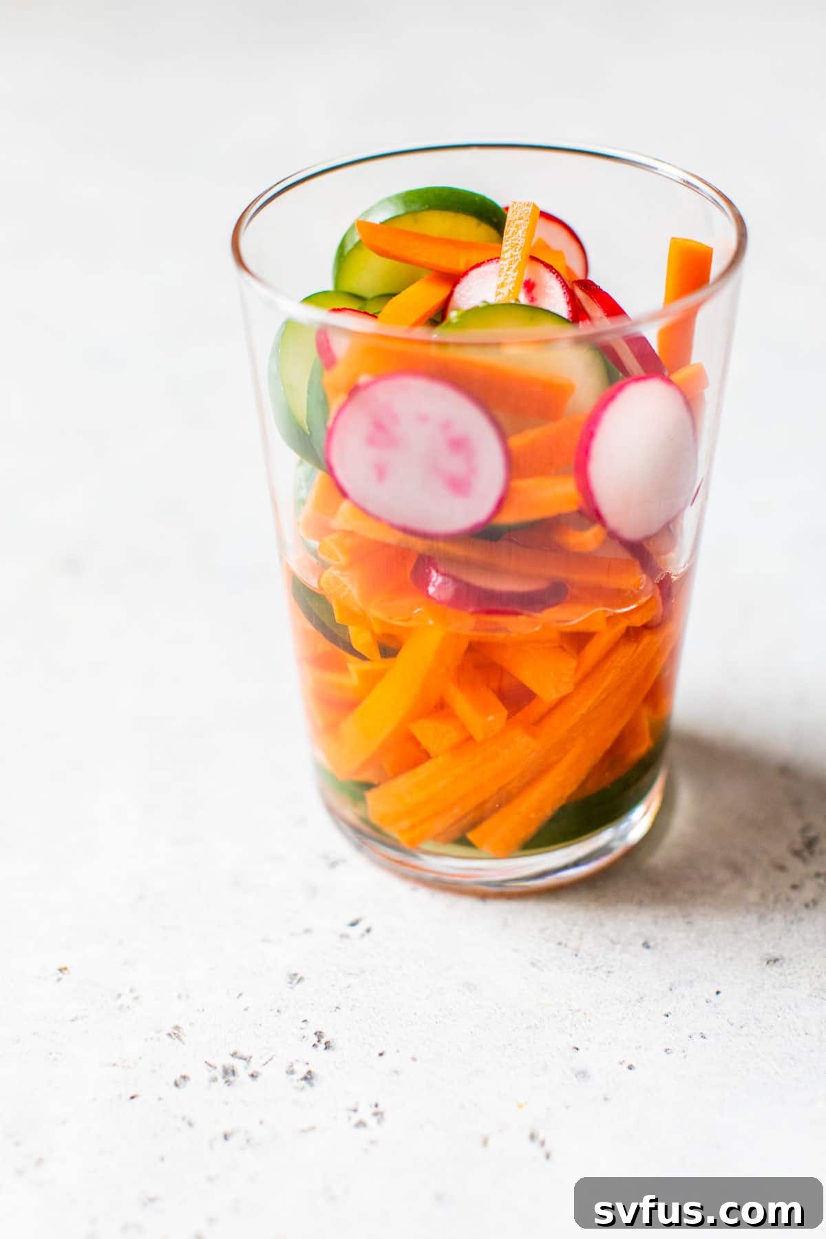 Close-up of thinly sliced vegetables, including carrots, cucumbers, and radishes, soaking in vinegar within a glass jar, ready for pickling.