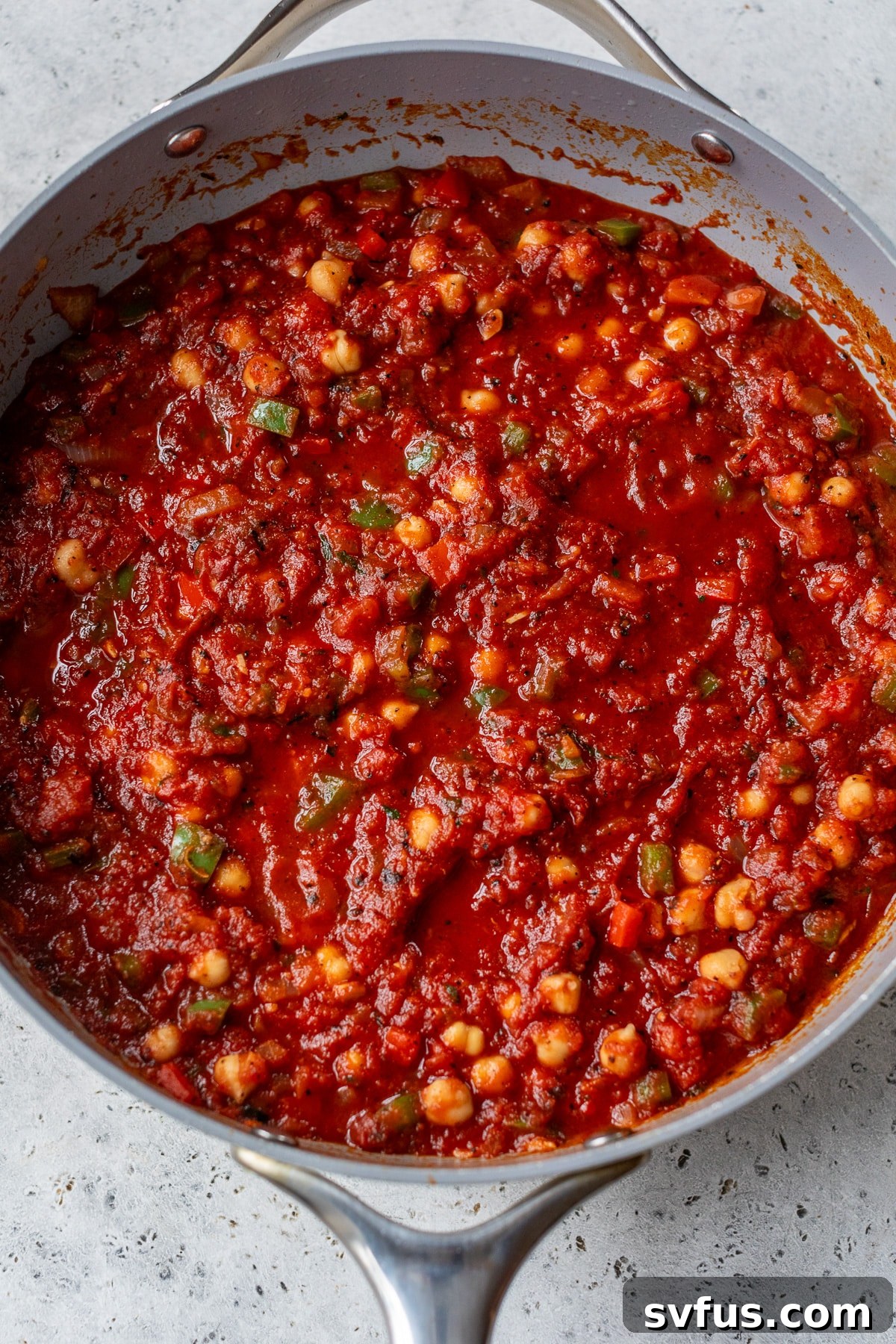 Crushed tomatoes and chickpeas being added to a skillet with sautéed vegetables and spices, simmering together to develop deep, rich flavors for vegan shakshuka