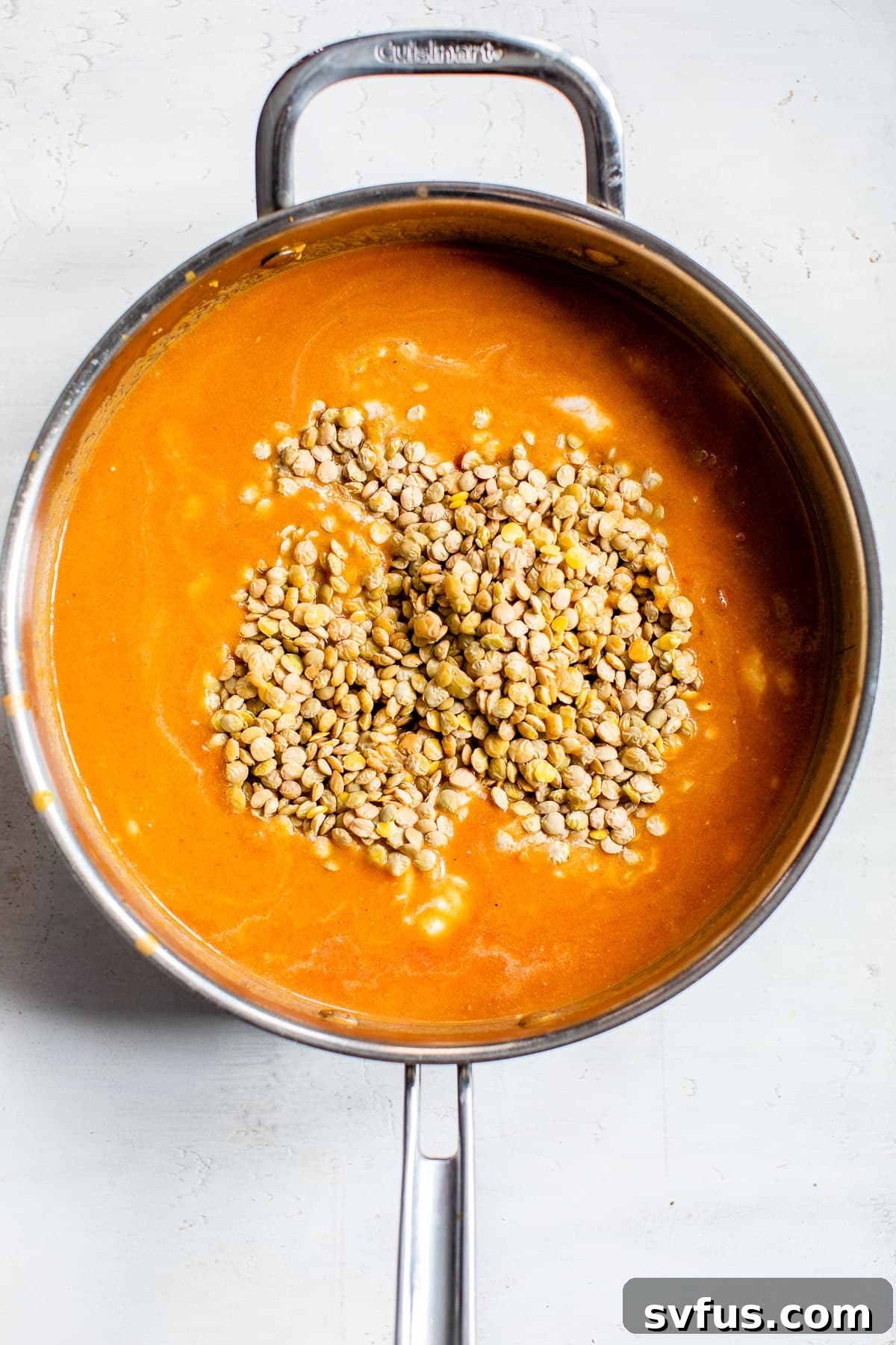 Uncooked lentils submerged in a vibrant orange-colored curry sauce, simmering in a skillet on the stovetop.