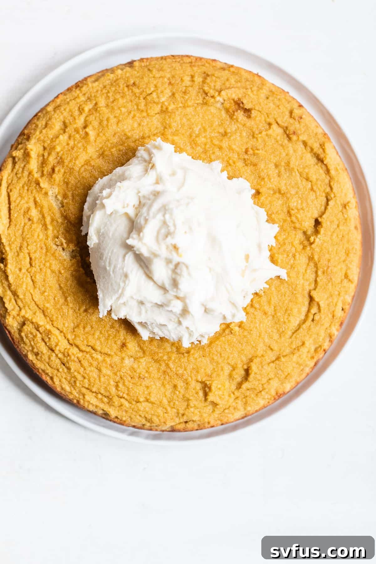 A freshly baked round cake, golden brown and perfectly cooled, with a generous dollop of creamy icing artfully placed on top, signaling readiness for spreading.