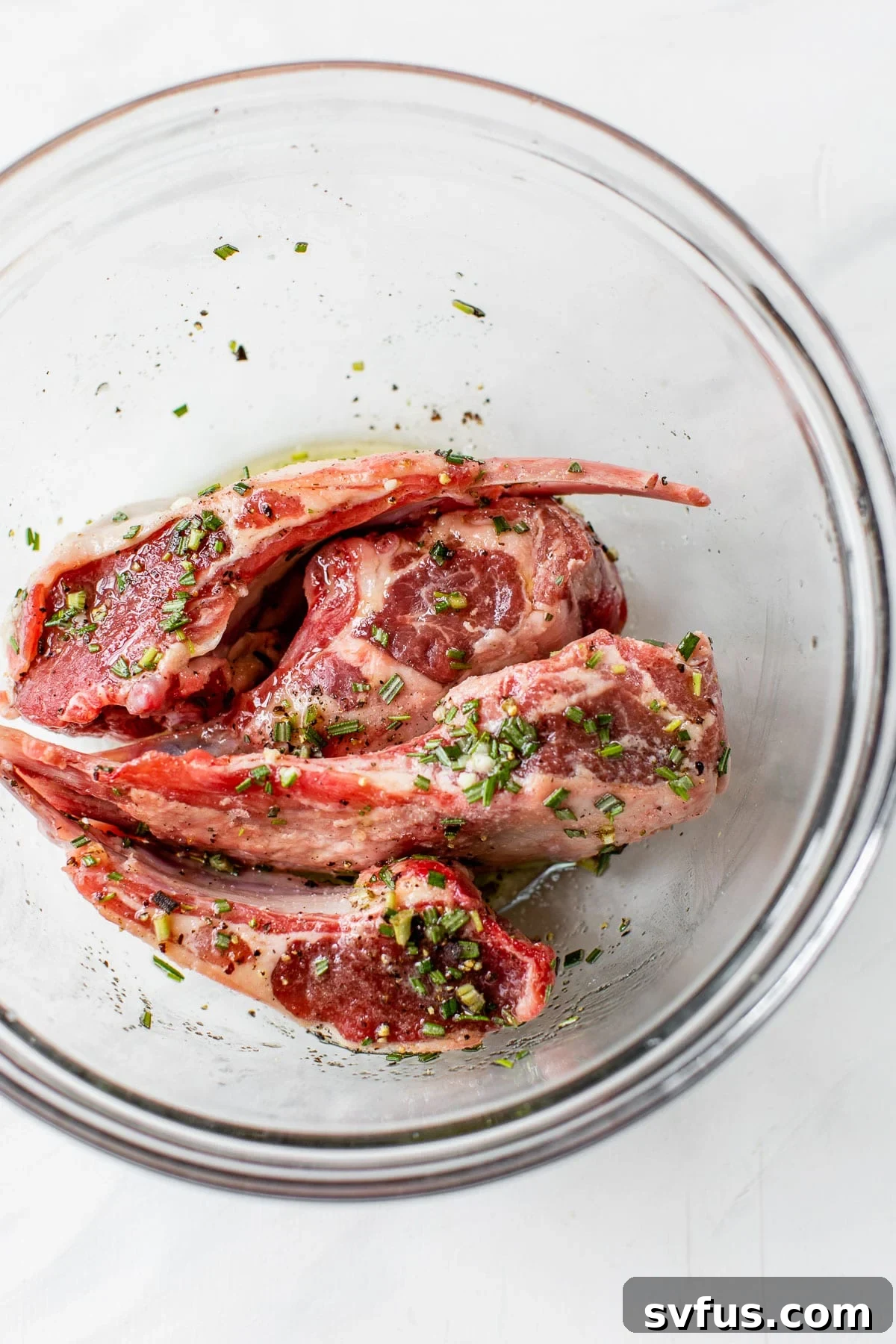 Raw lamb chops marinating in a clear glass bowl, coated in olive oil, minced garlic, and fresh rosemary.