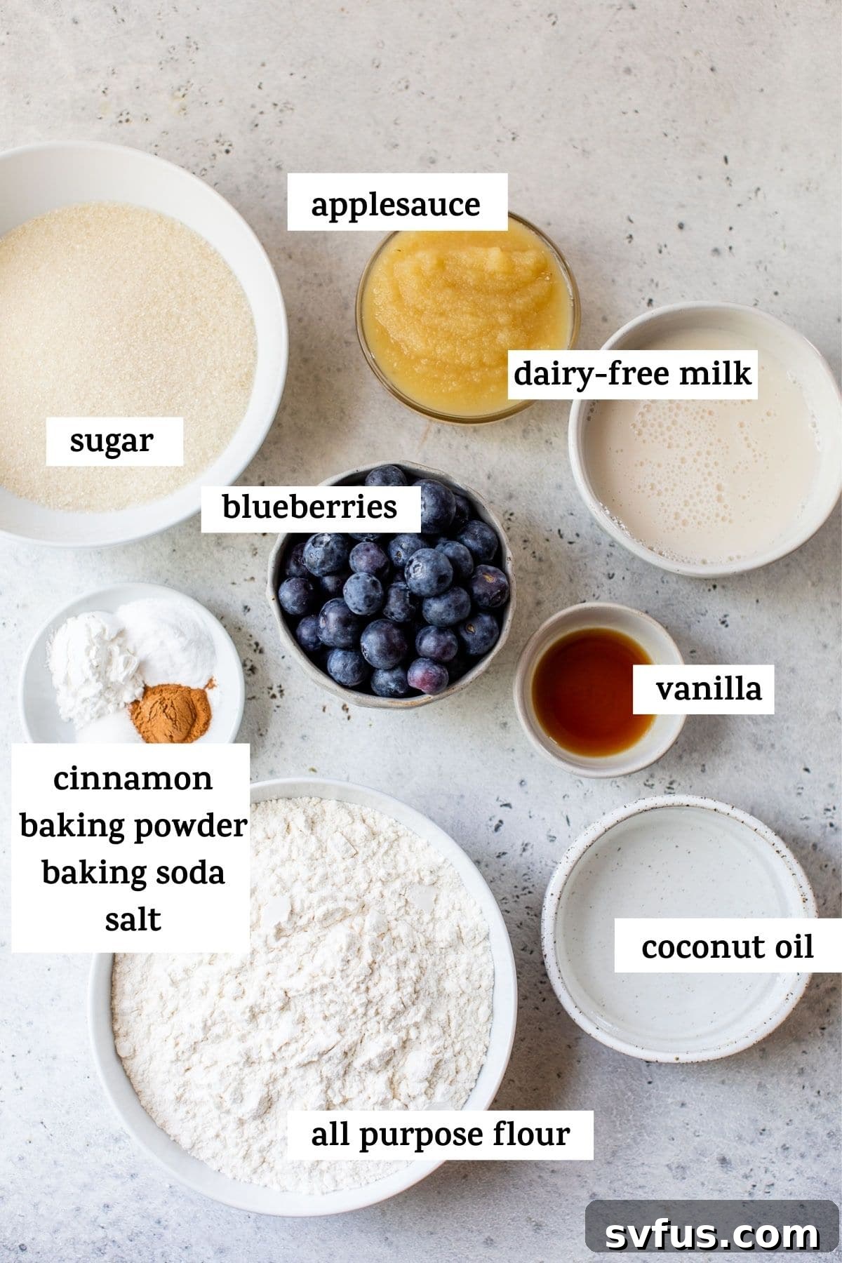 Assorted ingredients for making vegan blueberry muffins, including flour, sugar, applesauce, and fresh blueberries, laid out on a kitchen counter.