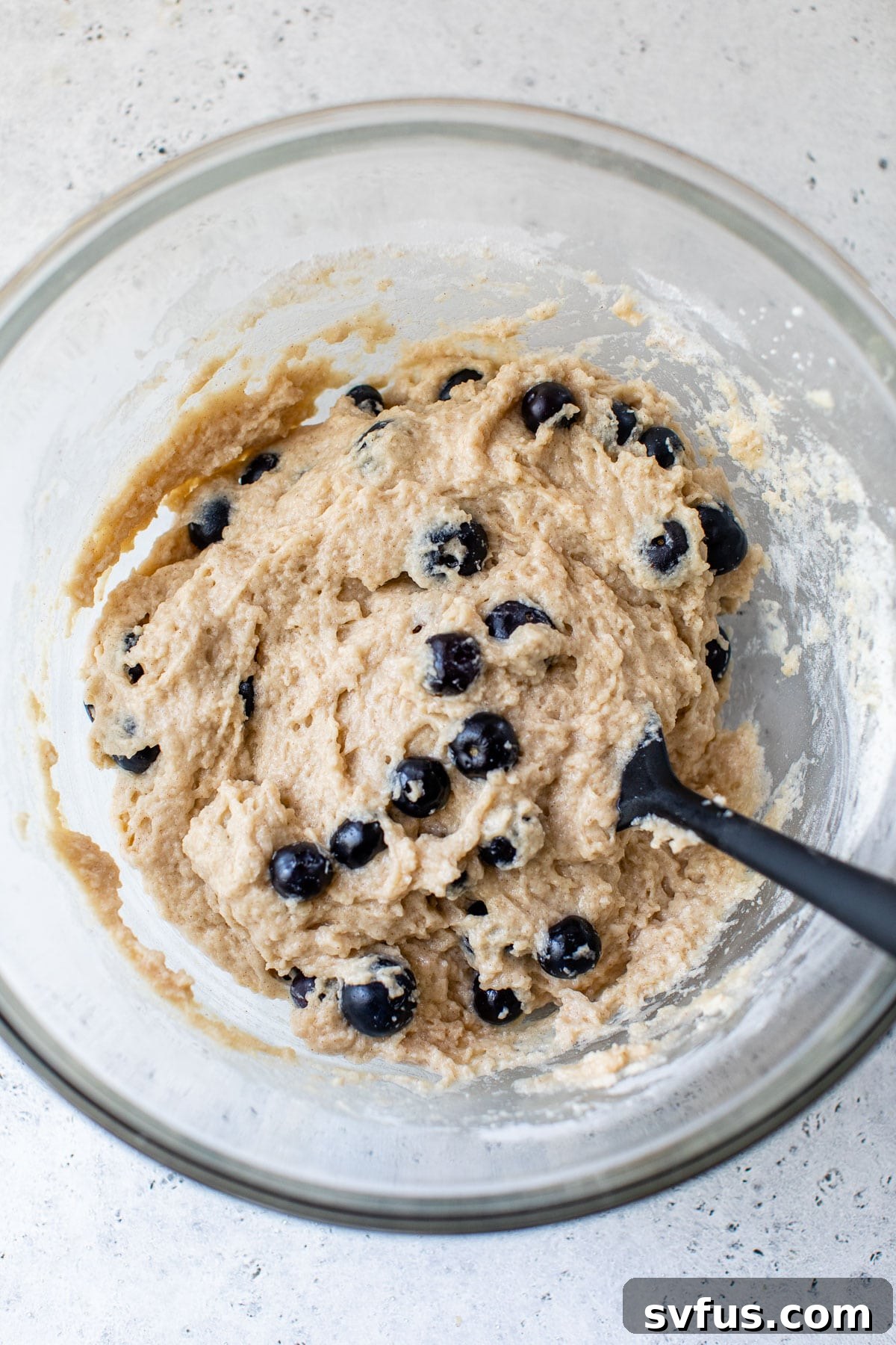 Thick vegan blueberry muffin batter in a large clear glass bowl, showcasing the blueberries mixed in.