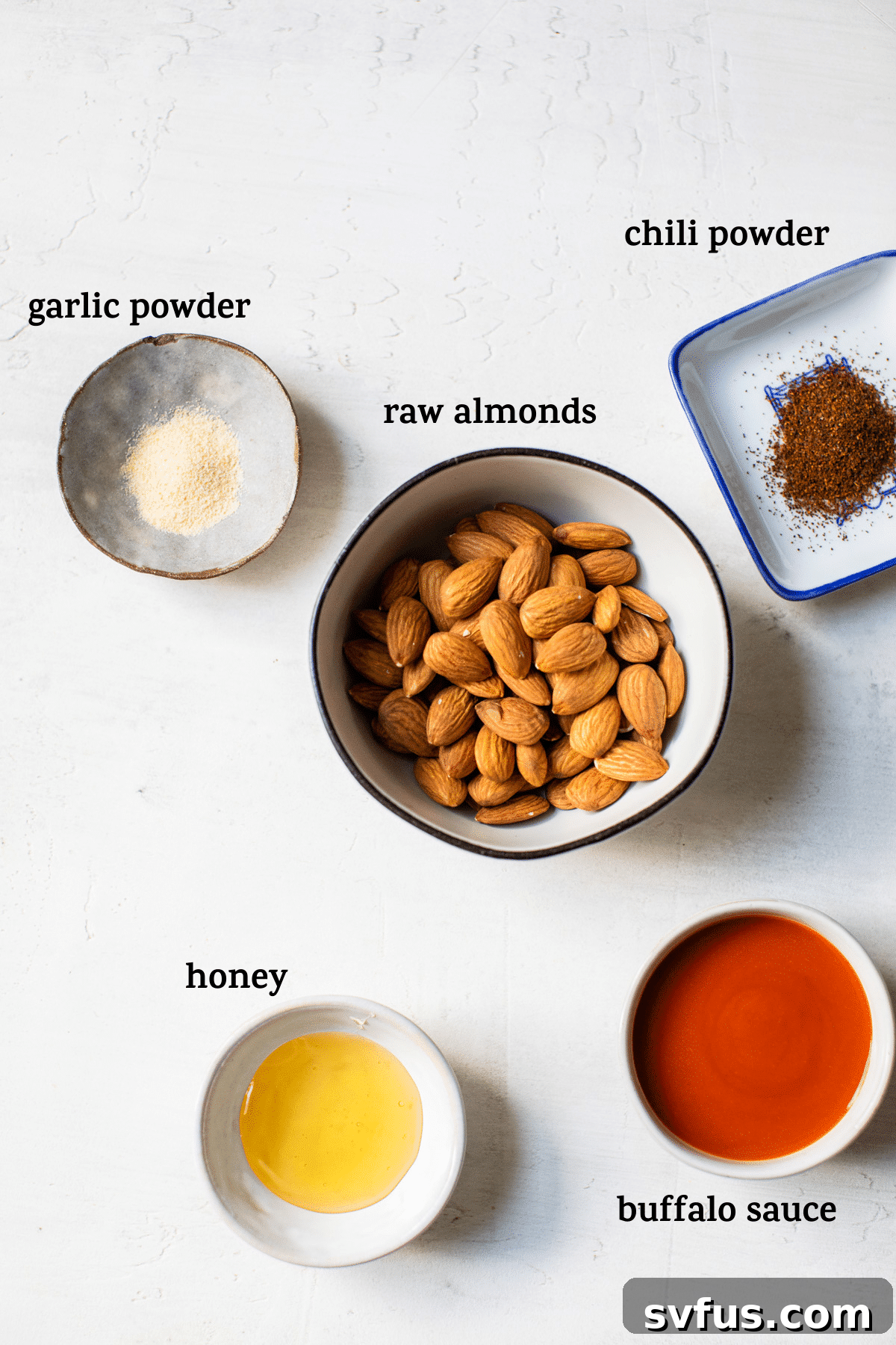 A collection of ingredients for buffalo almonds arranged on a wooden table, including raw almonds, a bottle of hot sauce, honey, chili powder, and garlic powder.
