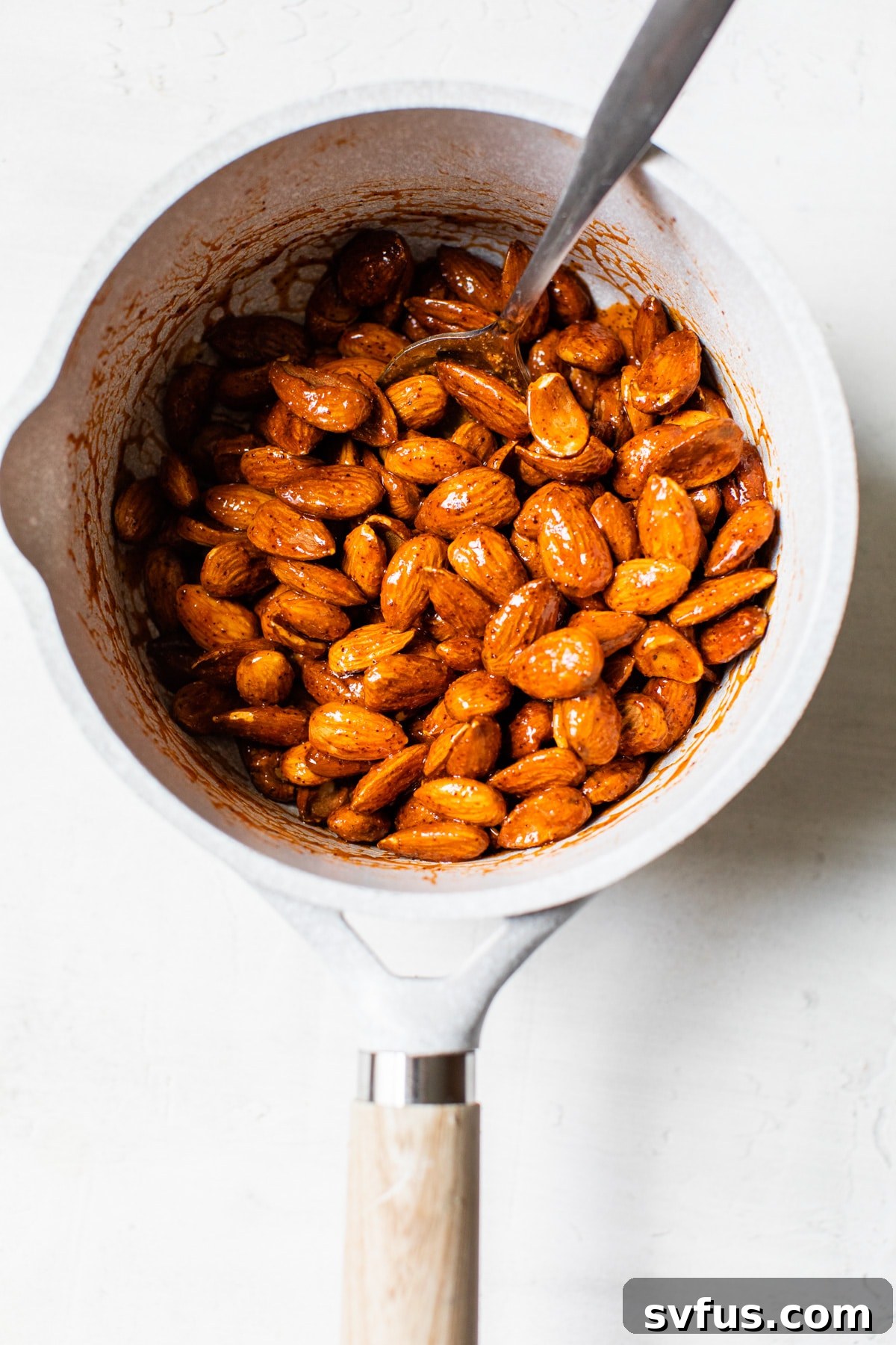 Raw almonds being stirred in a saucepan, completely coated in the reddish-orange buffalo sauce, ready for baking.
