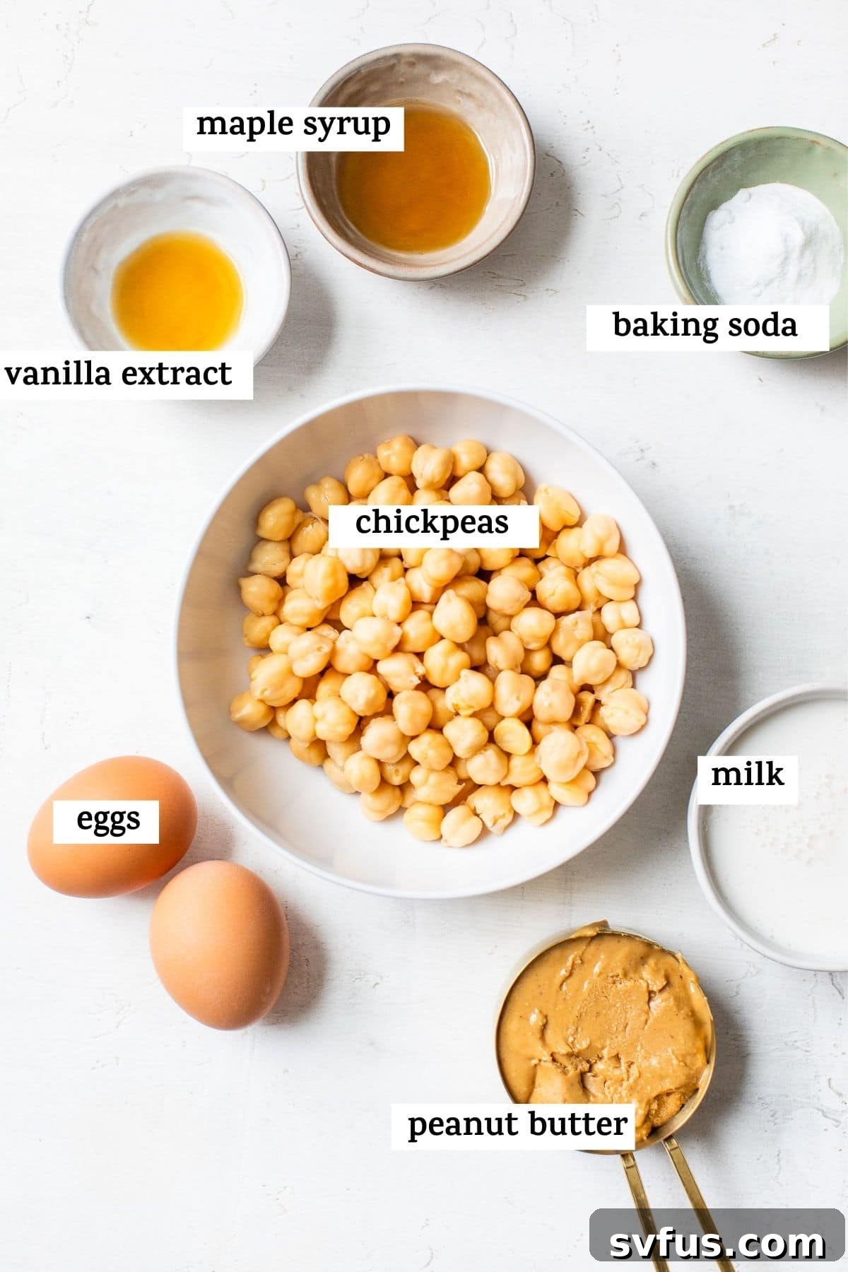 A collection of fresh ingredients laid out in bowls: chickpeas, eggs, peanut butter, baking soda, milk, vanilla extract, and maple syrup, ready for blending into pancake batter.