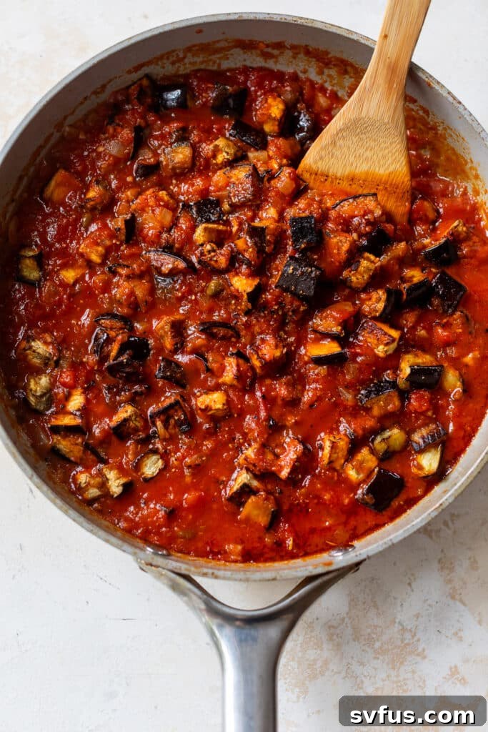 using a wooden spoon to stir the rich tomato sauce for eggplant pasta in a large skillet, adding depth to the flavors.