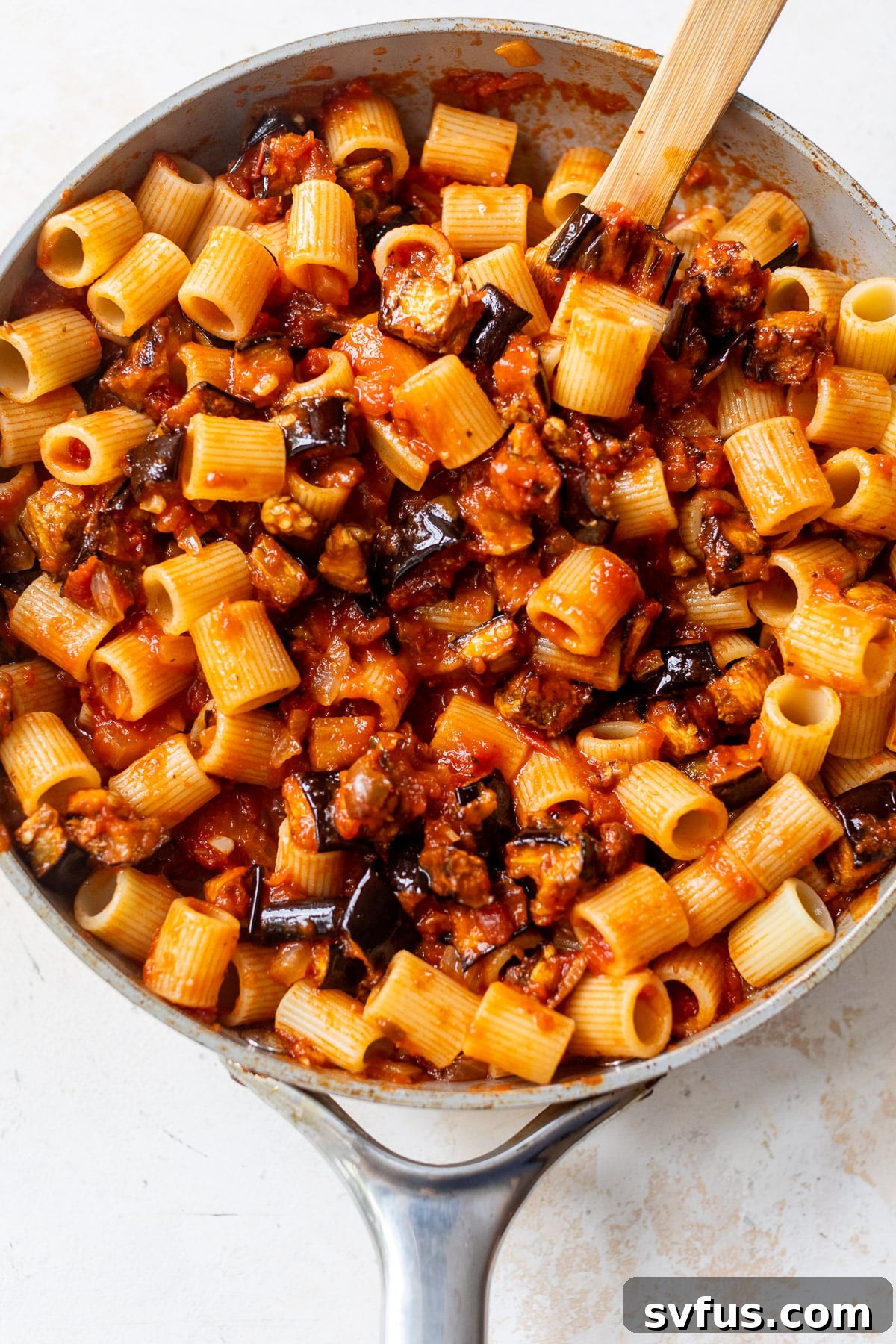 using a wooden spoon to stir the finished eggplant pasta in a large skillet, coated in savory tomato sauce.