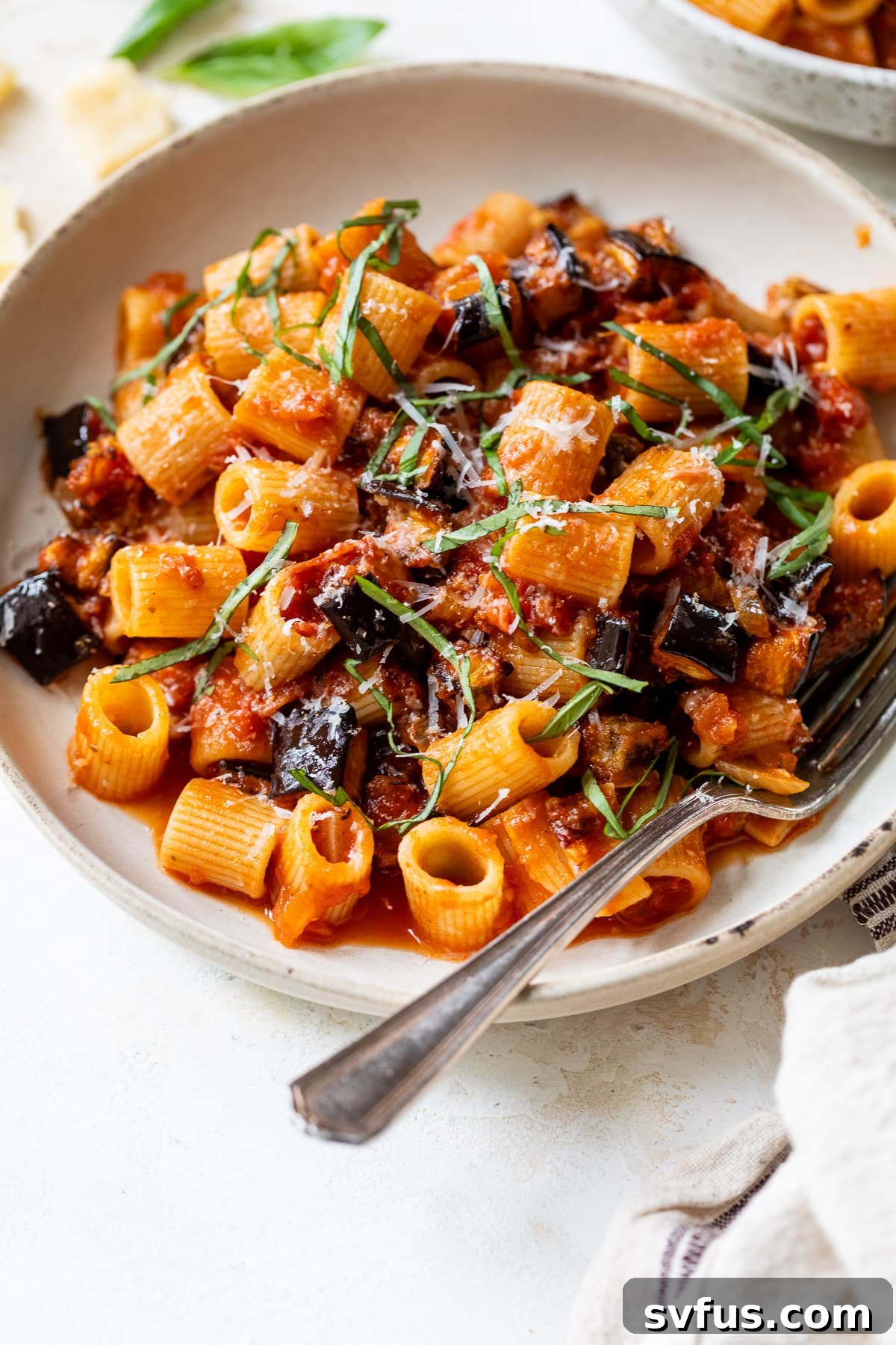 close up on a bowl of eggplant pasta topped with vibrant sliced basil and freshly grated parmesan cheese, ready to be enjoyed.