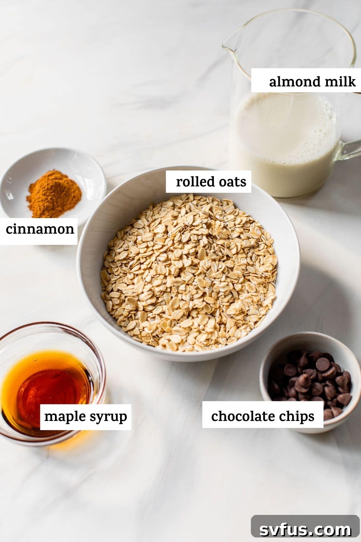 A selection of ingredients laid out on a table, including a bag of rolled oats, a carton of almond milk, a bottle of maple syrup, a jar of cinnamon, vanilla extract, and a bowl of chocolate chips, ready to be mixed into overnight oats.