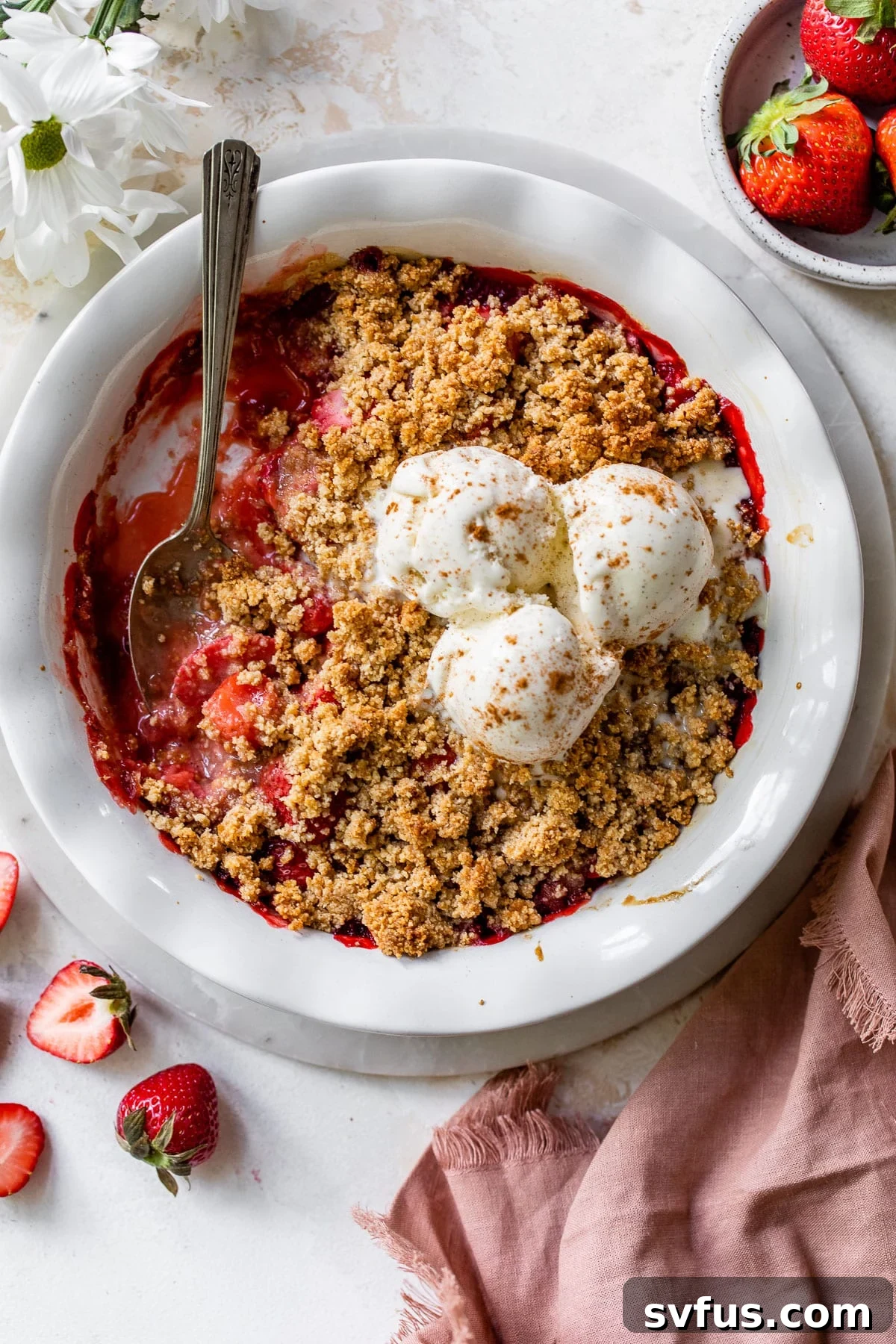 A beautifully baked strawberry crisp in a white pie pan, generously topped with three scoops of creamy vanilla ice cream, ready to be served.