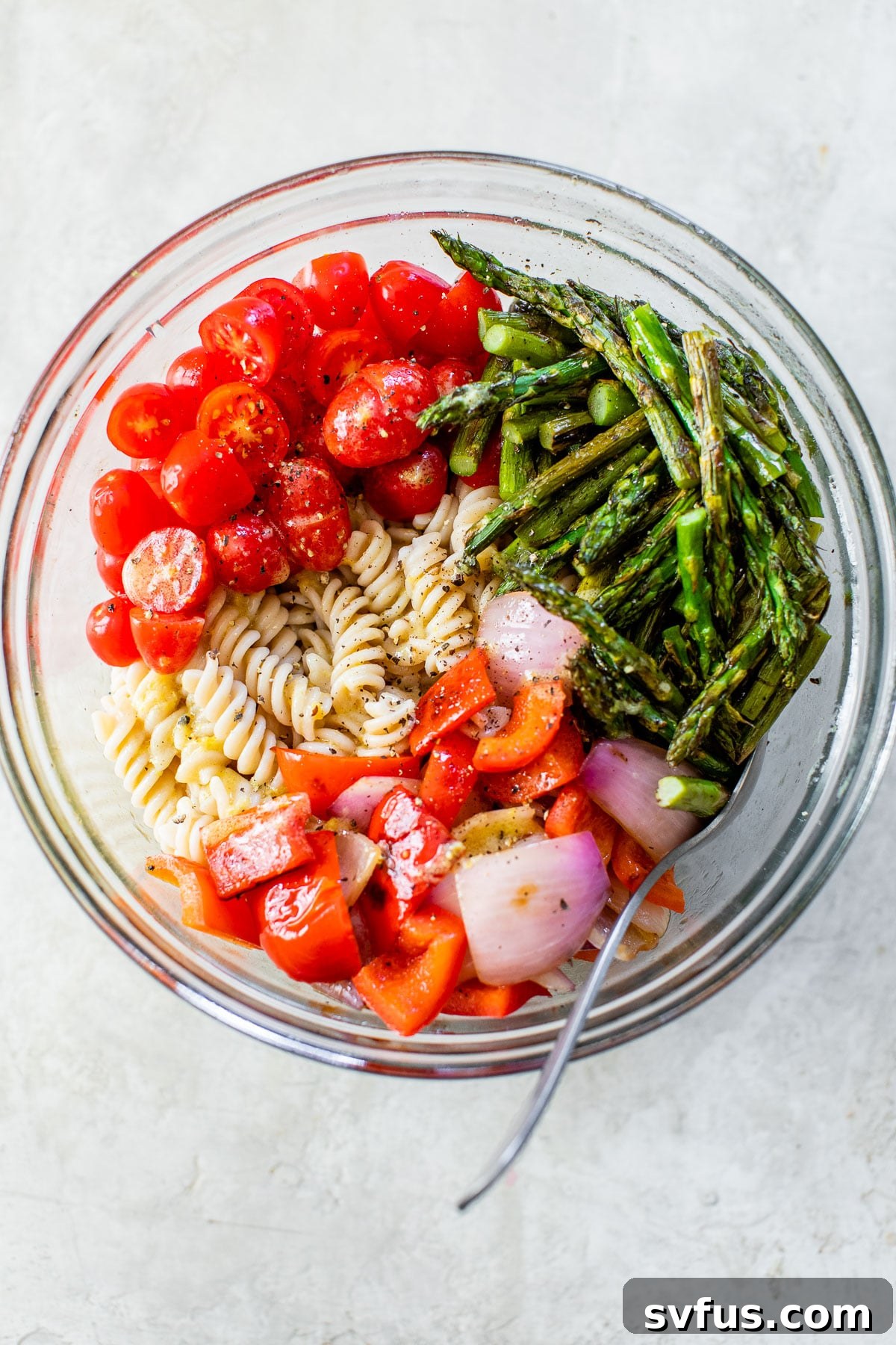 Close-up of cooked rotini pasta mixed with vibrant grilled asparagus, diced bell pepper, red onion, and fresh cherry tomatoes in a large serving bowl, ready to be dressed.