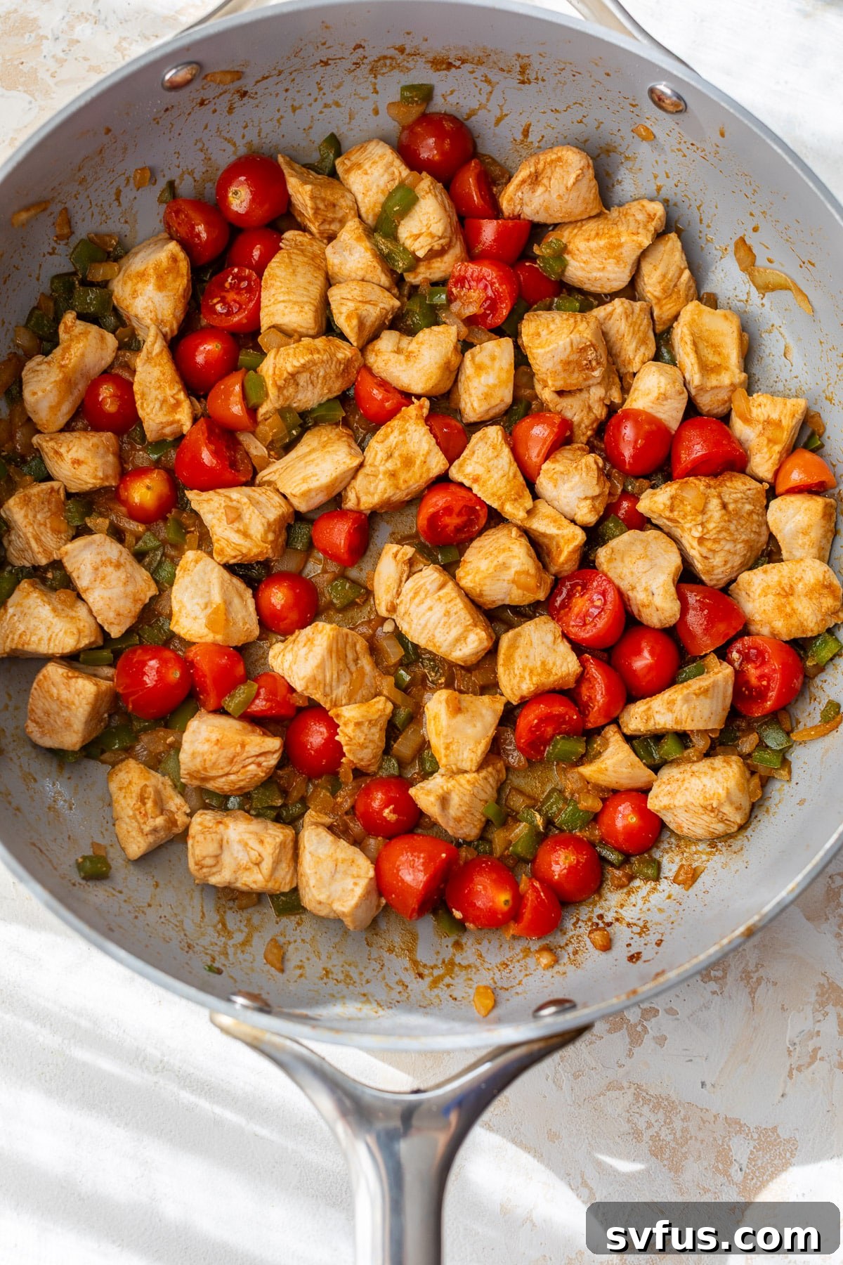 Close-up of diced chicken, bell peppers, onions, and grape tomatoes sizzling in a large skillet, perfectly seasoned with fajita spices.
