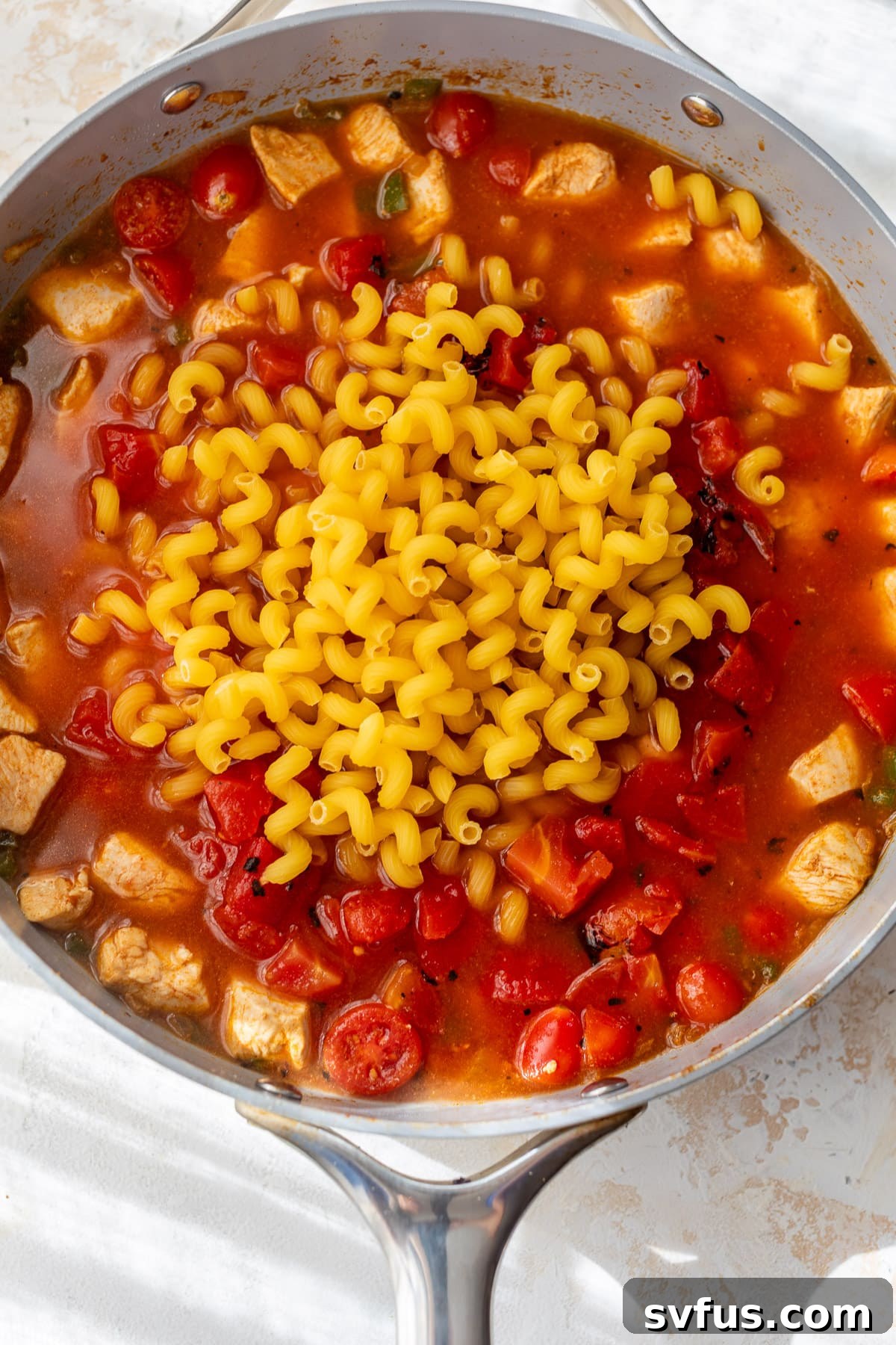 Uncooked short-cut pasta noodles submerged in a rich tomato sauce with cooked chicken pieces and vegetables in a large skillet, ready to simmer.