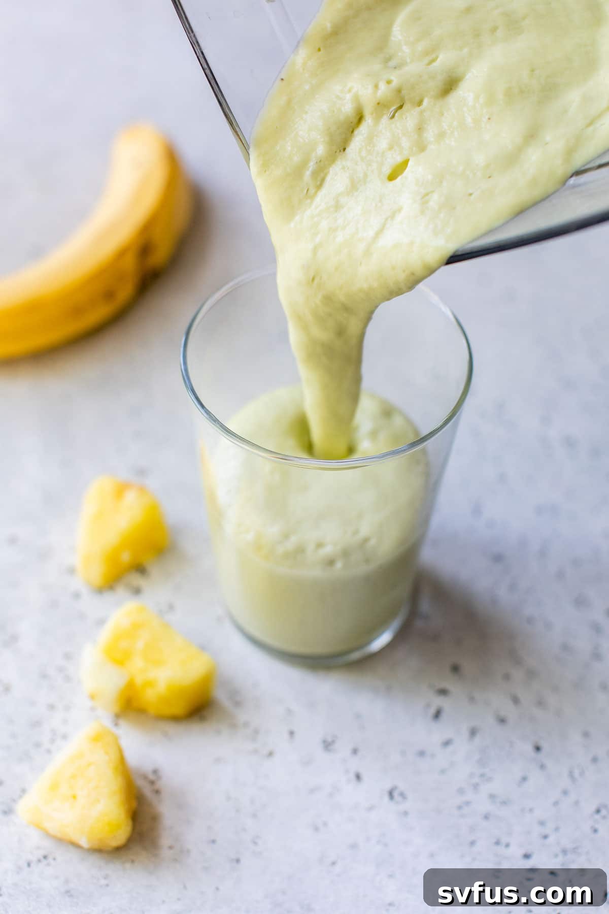 A smooth, green pineapple avocado smoothie being poured from a blender into a tall glass.