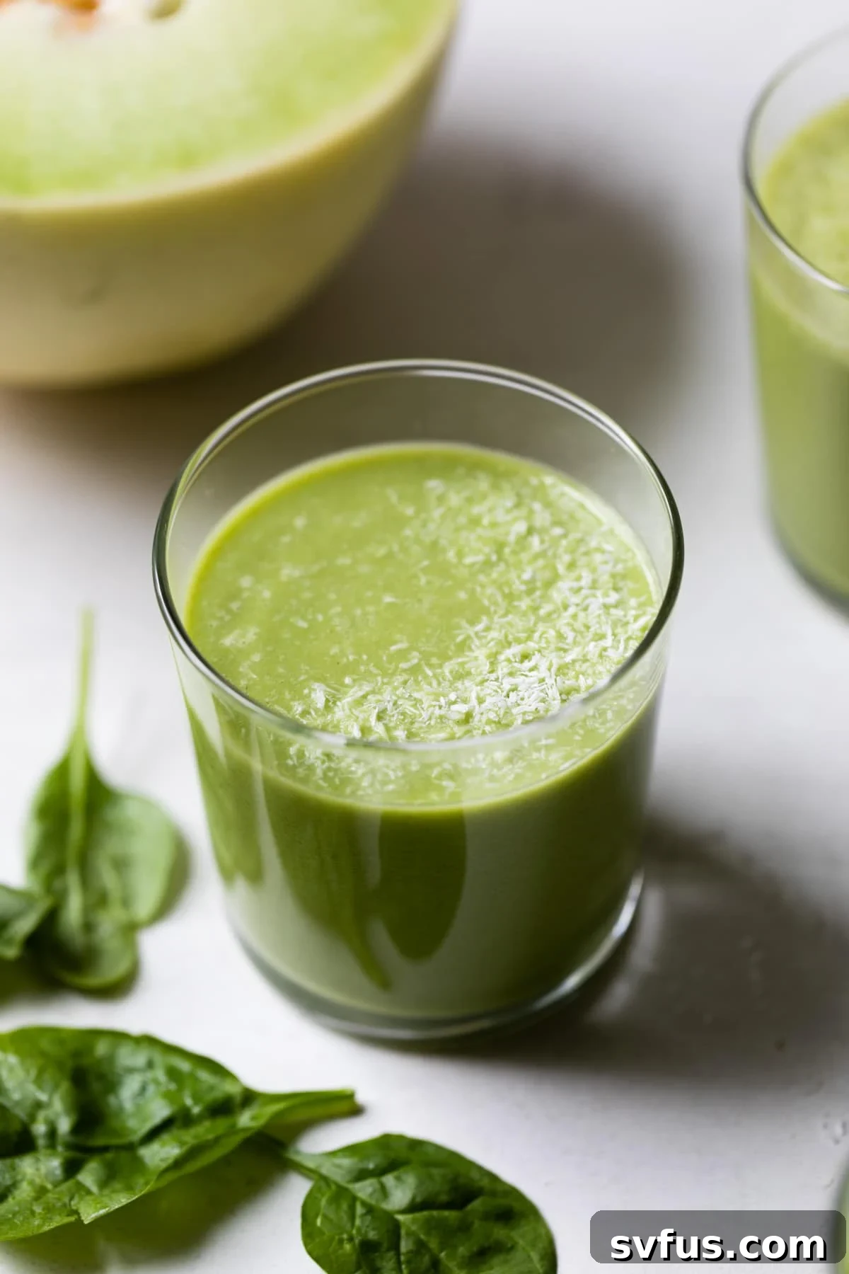 Vibrant green honeydew smoothie in a tall glass, garnished with a small honeydew wedge, against a light background.