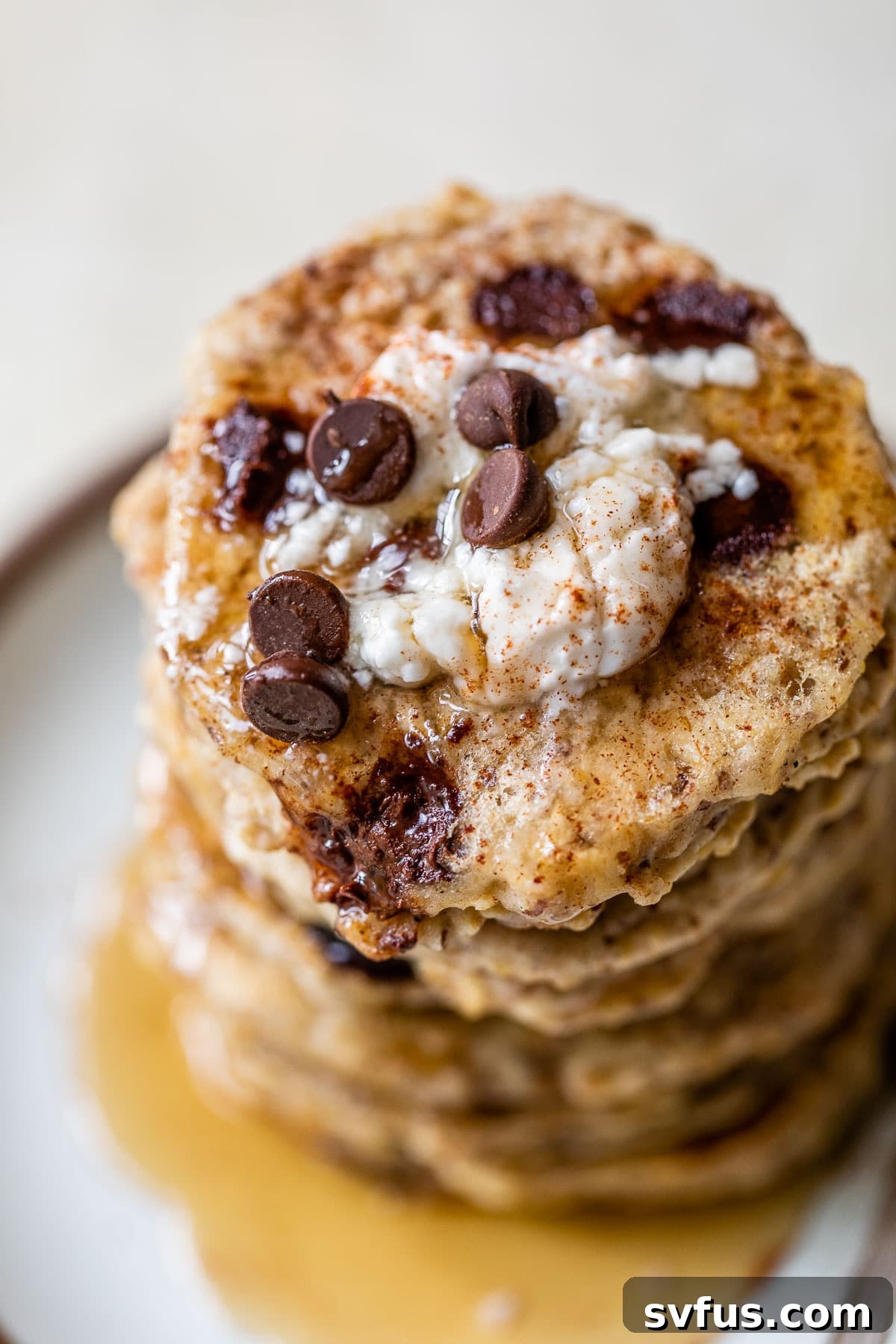 A tall stack of fluffy vegan oatmeal pancakes, generously topped with creamy vegan yogurt and a sprinkle of chocolate chips, photographed on a rustic plate.
