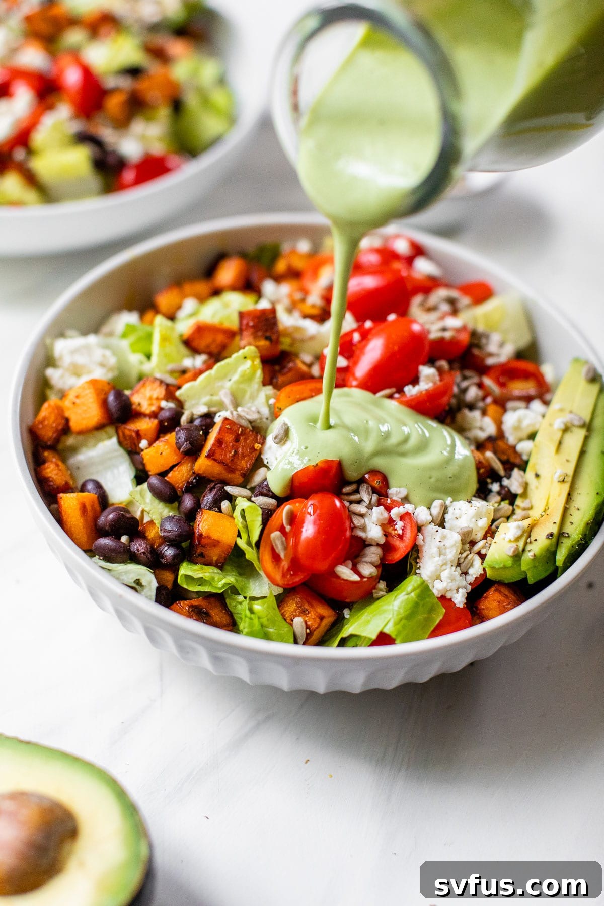 A hand pouring vibrant green spicy avocado dressing over a beautifully assembled sweet potato salad in a bowl.