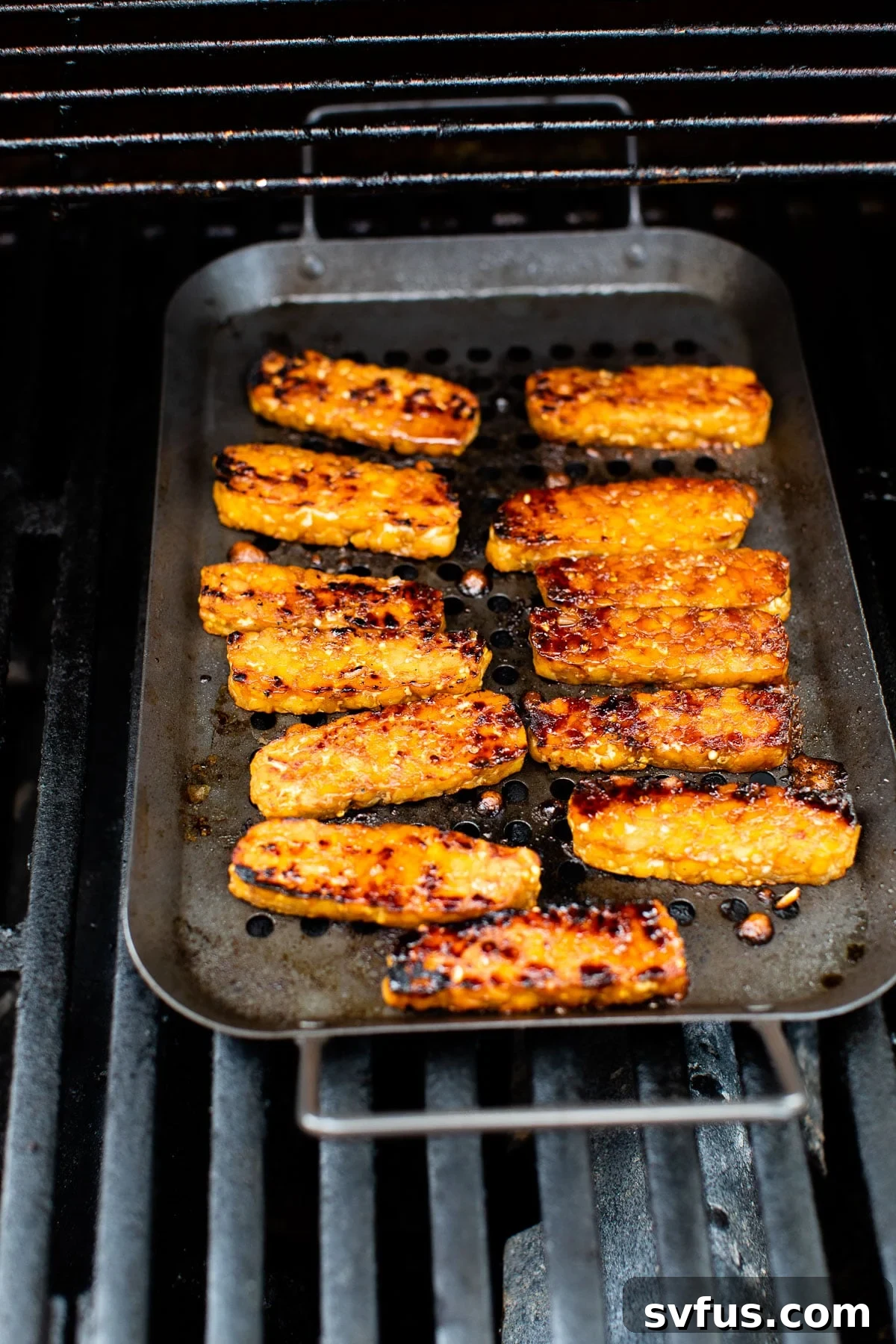 Marinated tempeh slices cooking on a hot outdoor grill grate, showing developing char marks.