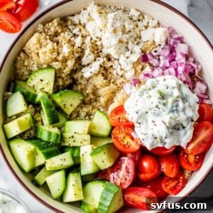 a bowl of quinoa topped with mediterranean ingredients like tomatoes, cucumber, red onion, and feta