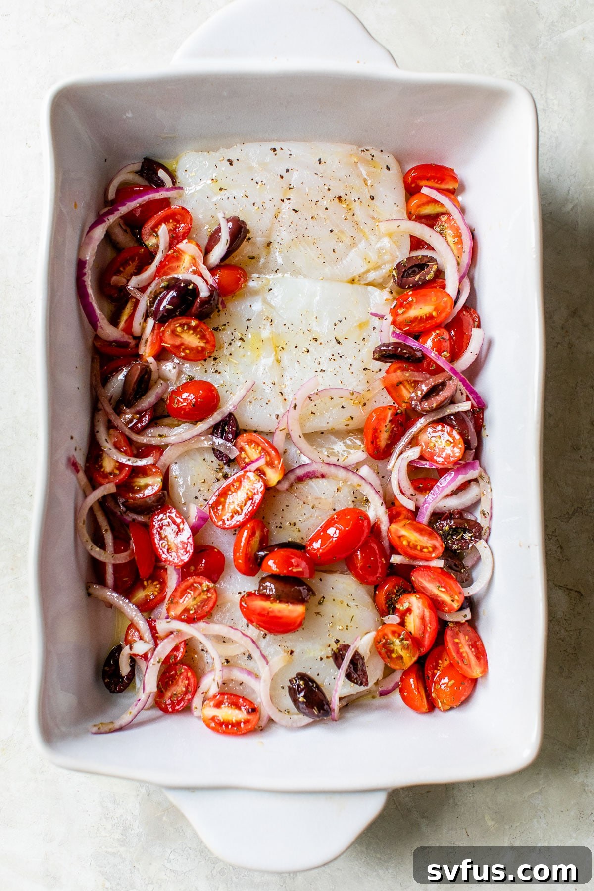 Raw cod fillets in a baking dish topped with grape tomatoes and other Mediterranean vegetables.