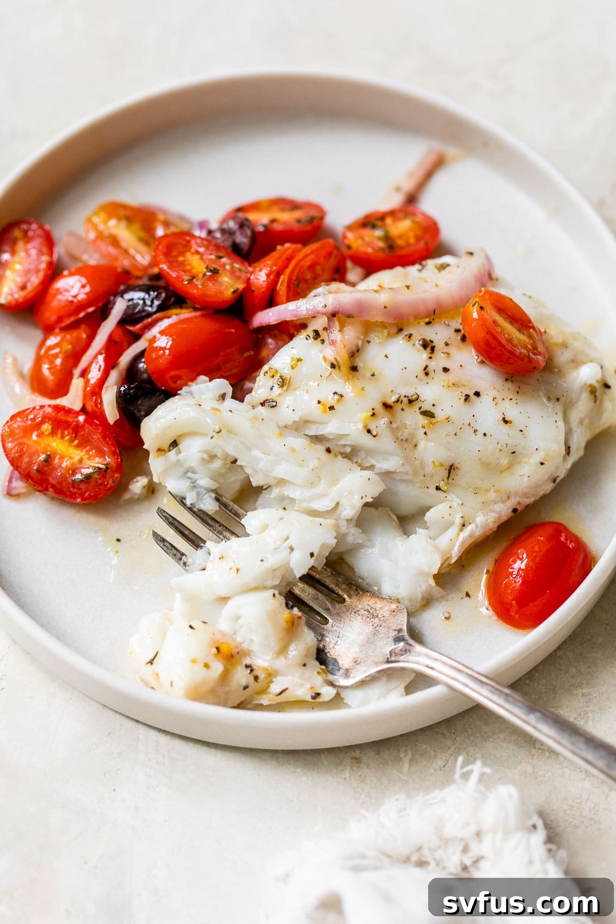 A single piece of white fish on a plate beside cooked grape tomatoes and herbs.