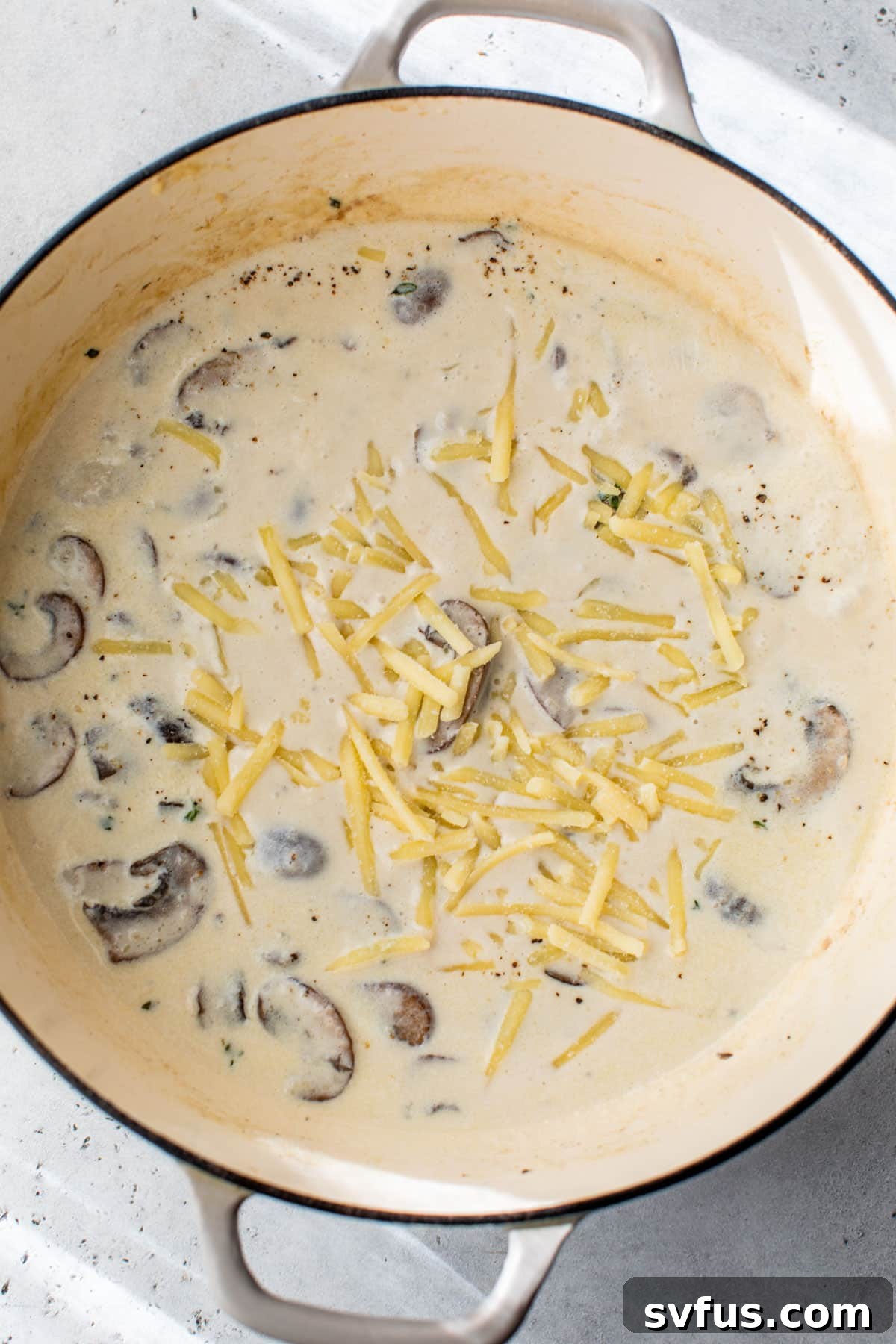 Shredded cheese being gradually added to the creamy sauce in a Dutch oven with mushrooms
