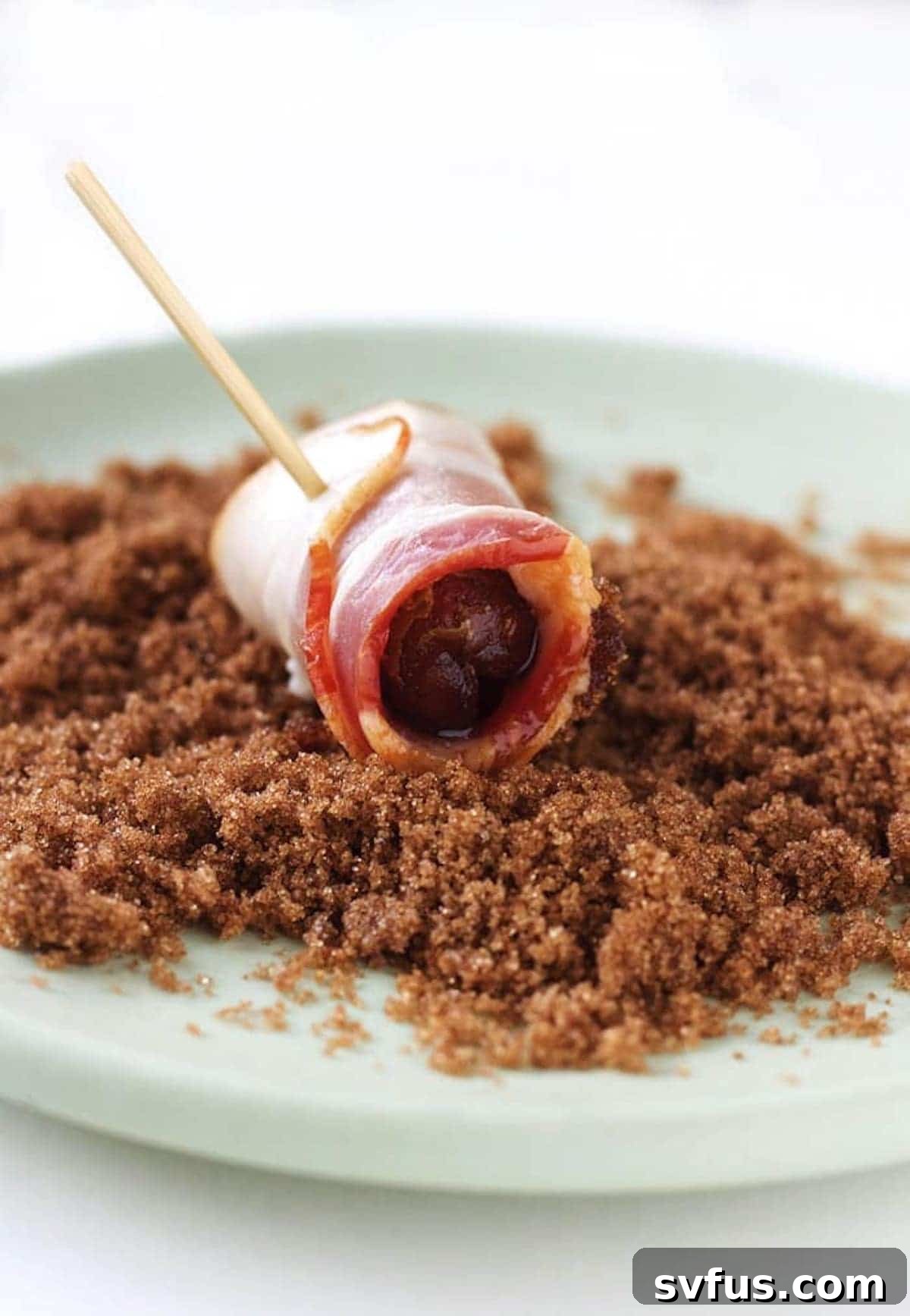 A hand dipping an uncooked bacon-wrapped date into a bowl of brown sugar before baking.