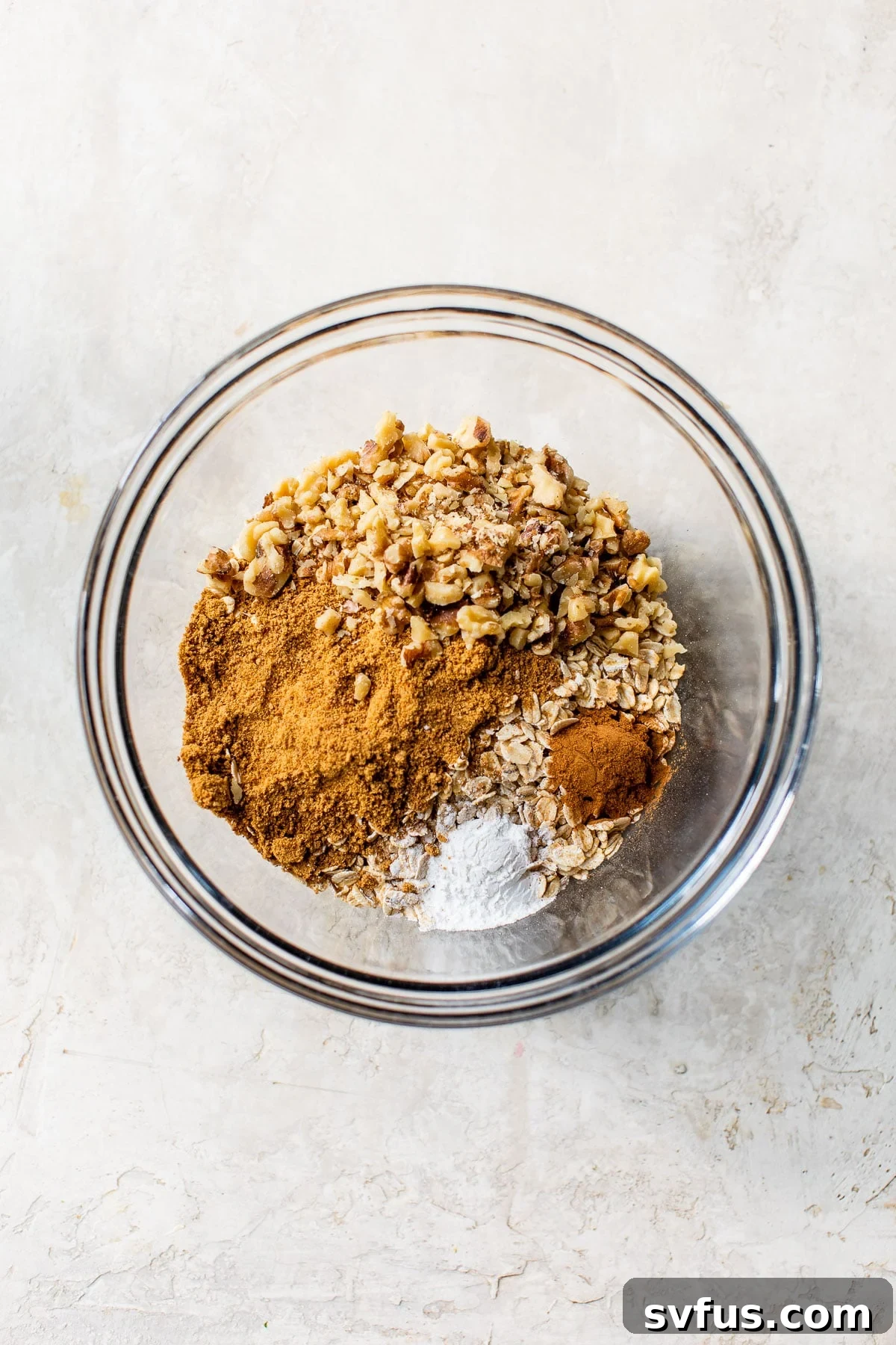 A large glass bowl filled with the dry ingredients for baked oatmeal: rolled oats, coconut sugar, cinnamon, baking powder, and walnuts, ready to be combined.