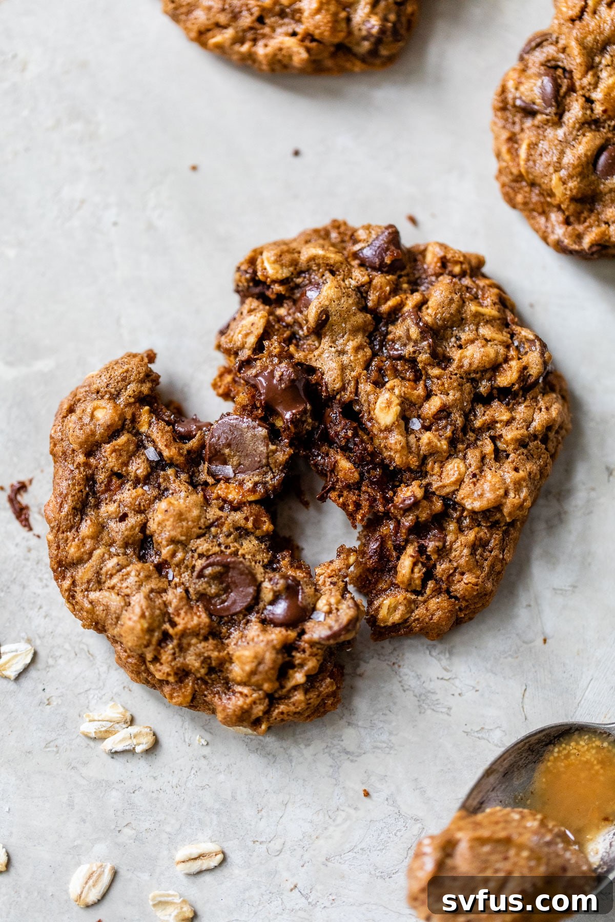 A freshly baked almond butter oatmeal cookie, artfully broken in half to reveal its soft, chewy interior texture and the abundance of oats and chocolate chips.